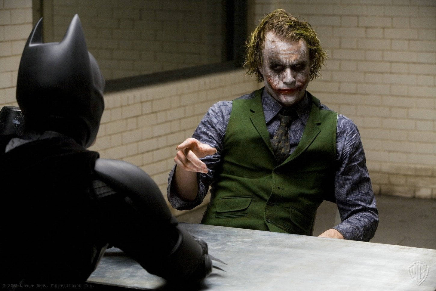 Heath Ledger Joker 桌布 壁紙 — 小丑穿著綠色背心，在一個簡陋的磚牆房間裡，隔著桌子與蝙蝠俠面對面。