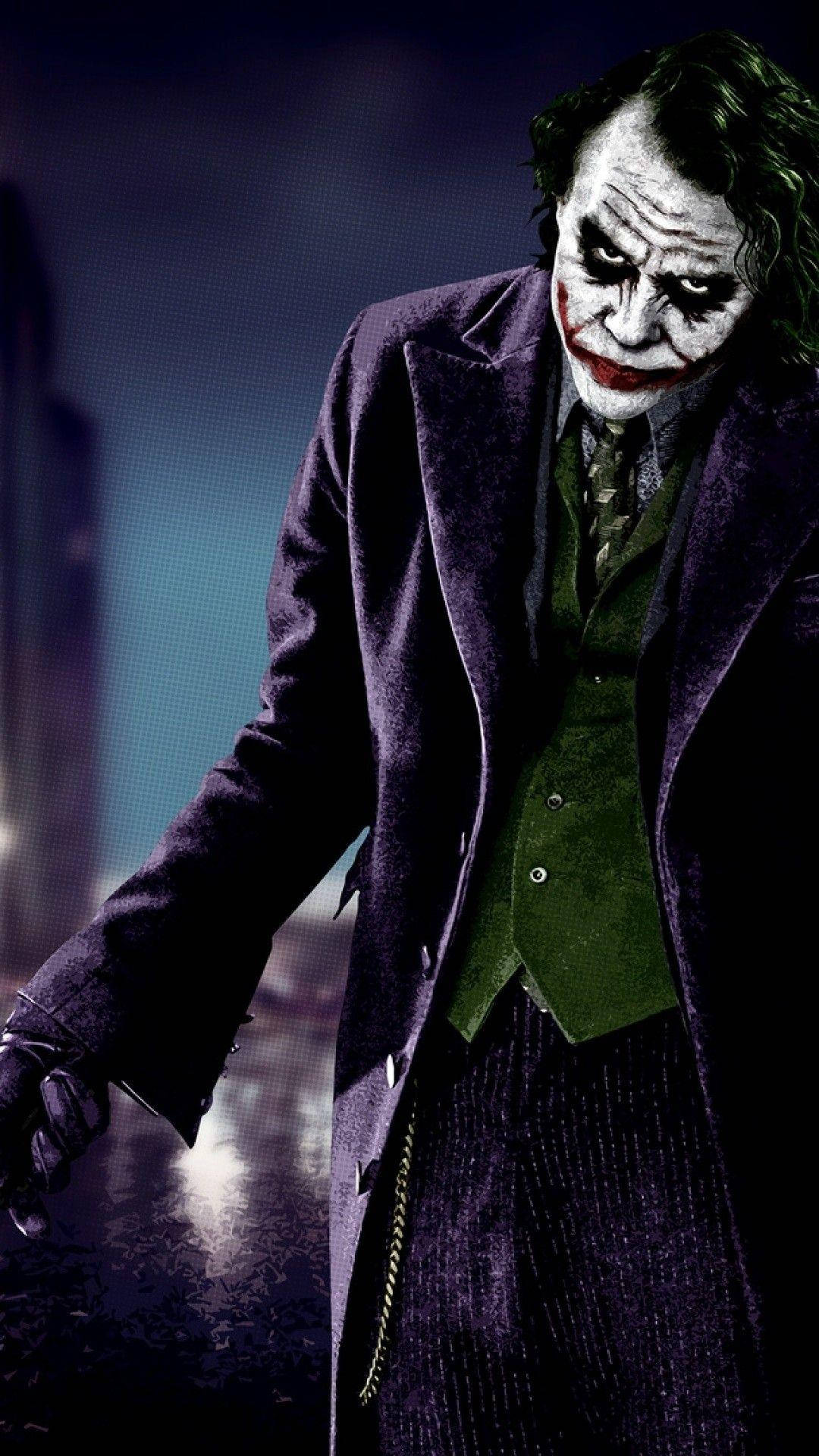 Heath Ledger 桌布 壁紙 — 小丑身穿紫色外套和綠色背心，在黑暗的城市背景下，帶著發光的燈光，威嚇地站立。