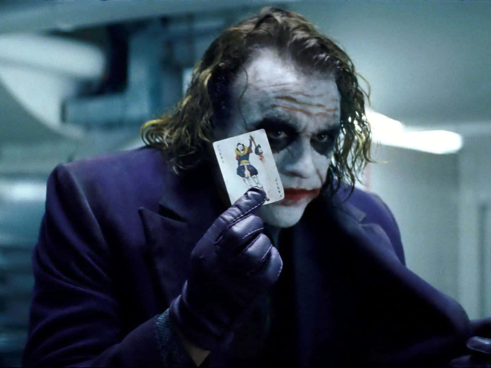 Heath Ledger Joker 桌布 壁紙 — 小丑身穿紫色西裝，在昏暗的工業環境中手持一張撲克牌，露出陰險的笑容。