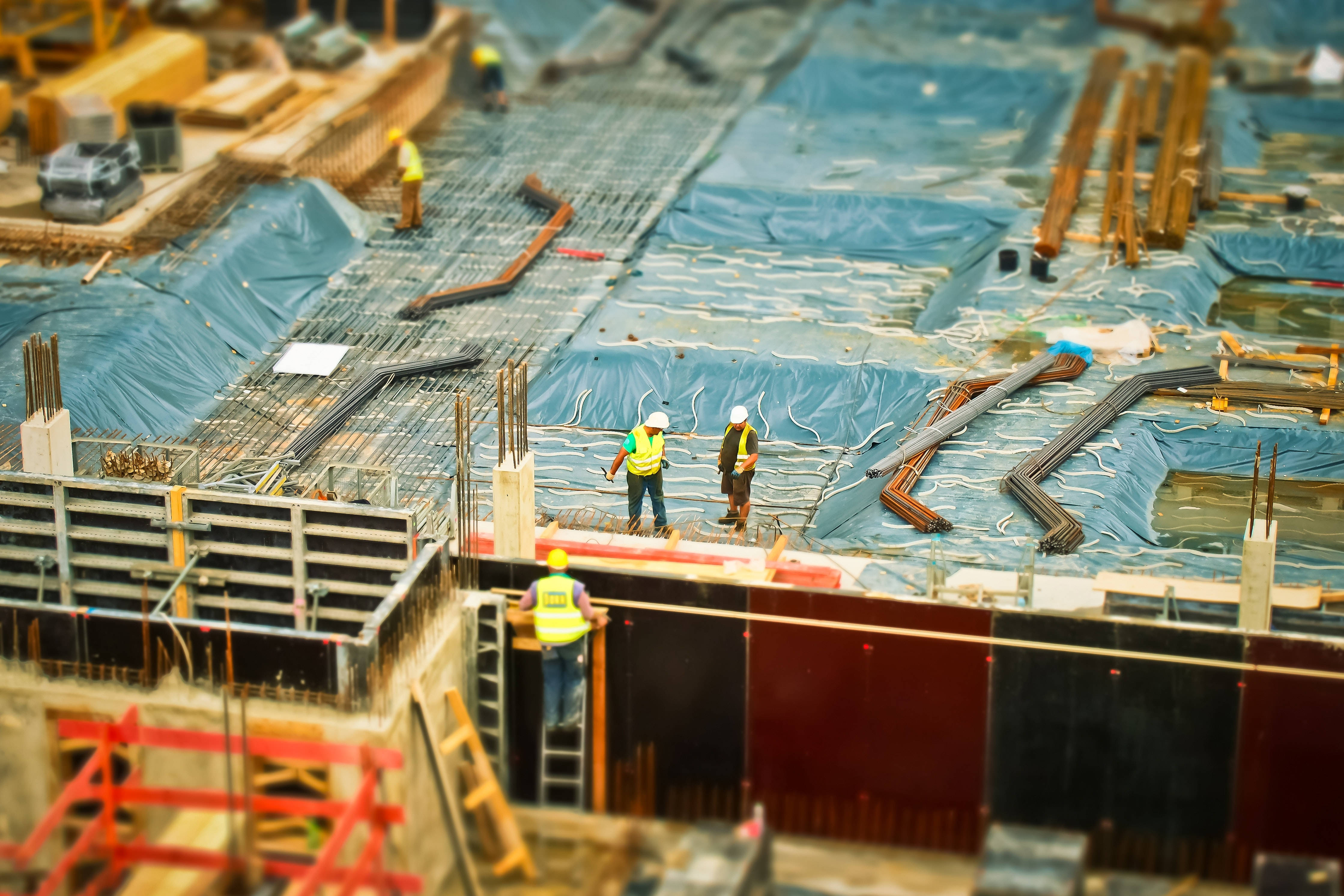 Working 桌布 壁紙 — 小小的建築工人穿著鮮豔背心，站在一個鋪著藍色防水布、有鋼筋和鷹架的廣闊工地上，在溫暖的日光下。