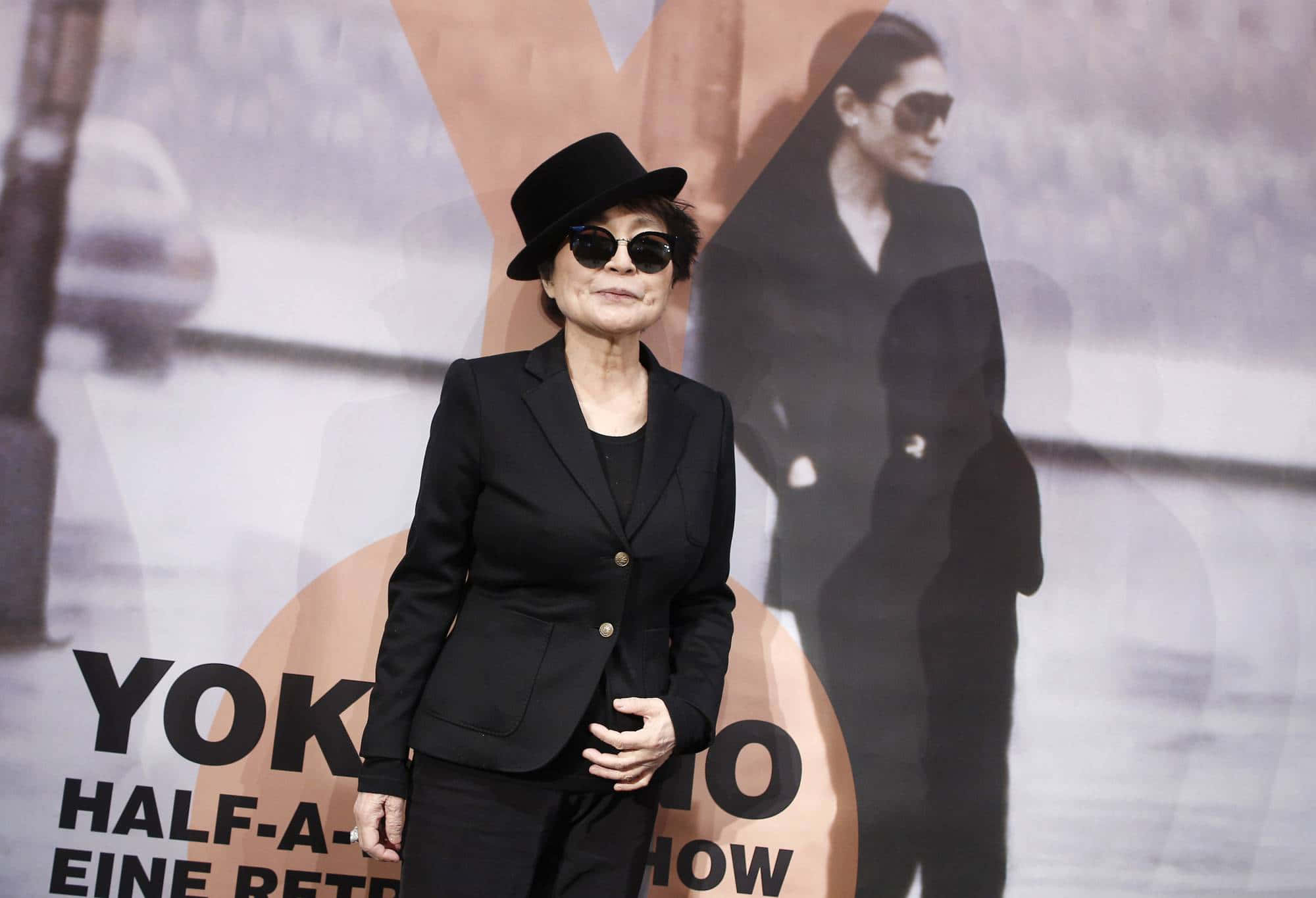 Sunglasses 桌布 壁紙 — 小野洋子自信地穿著黑色西裝和帽子，站在她的「Half-A-Life」展覽的宣傳背景前。