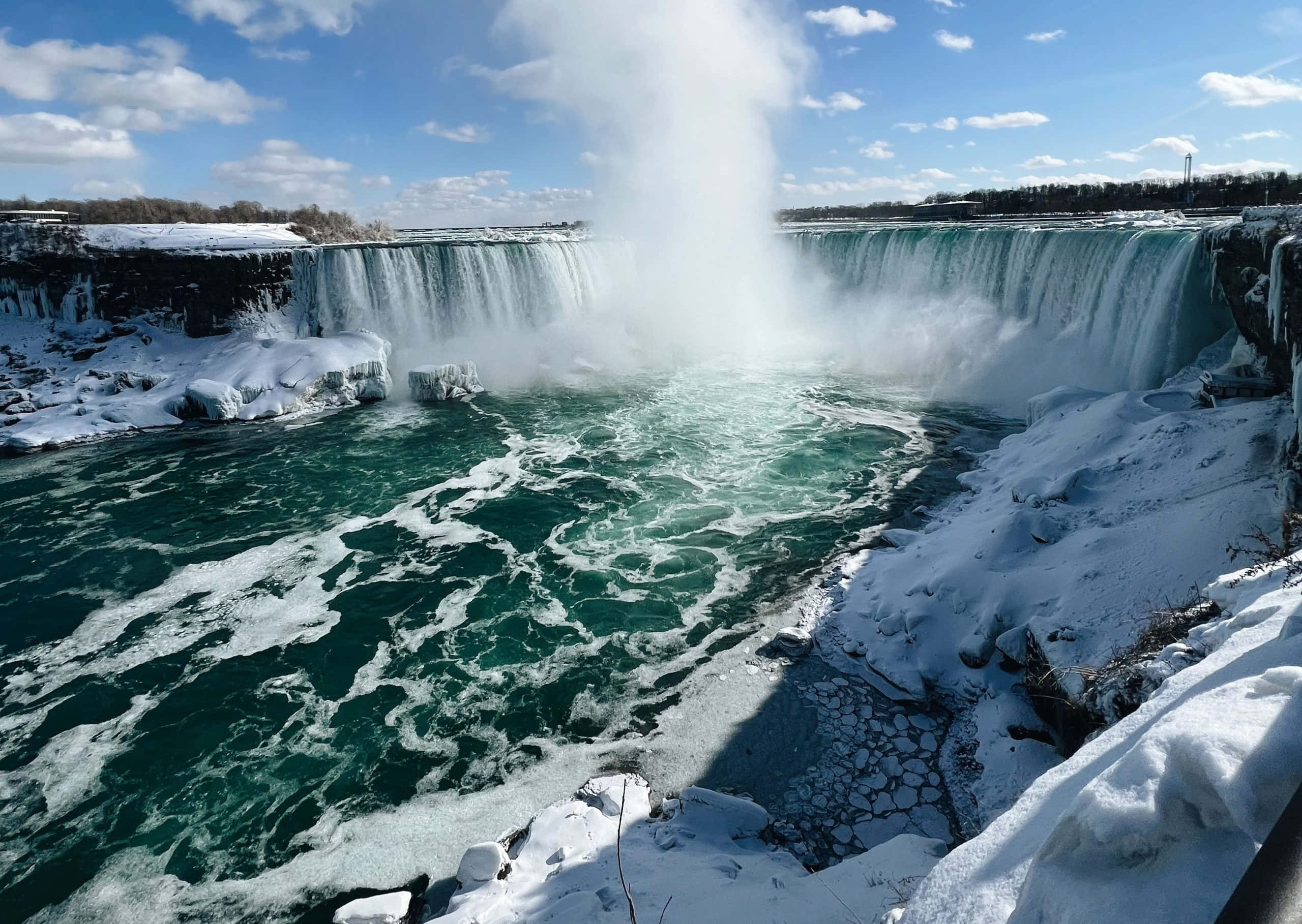 Niagara Falls 桌布 壁紙 — 尼亞加拉大瀑布在冬季強勁奔流，雪覆蓋的河岸和霧氣在明亮的藍天下升起。