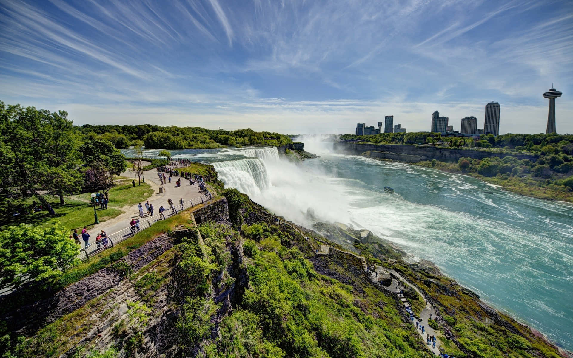720p Niagara Falls 桌布 壁紙 — 尼亞加拉大瀑布在明亮的藍天和薄雲下，周圍綠意盎然，水流奔騰而下的鳥瞰圖。