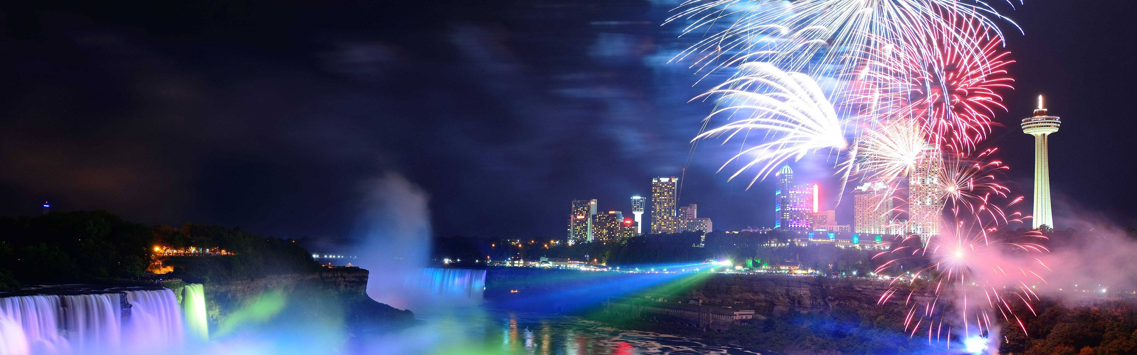 Fireworks 桌布 壁紙 — 尼亞加拉大瀑布在璀璨燈光下閃耀，城市天際線上綻放絢麗煙花的壯麗夜景。