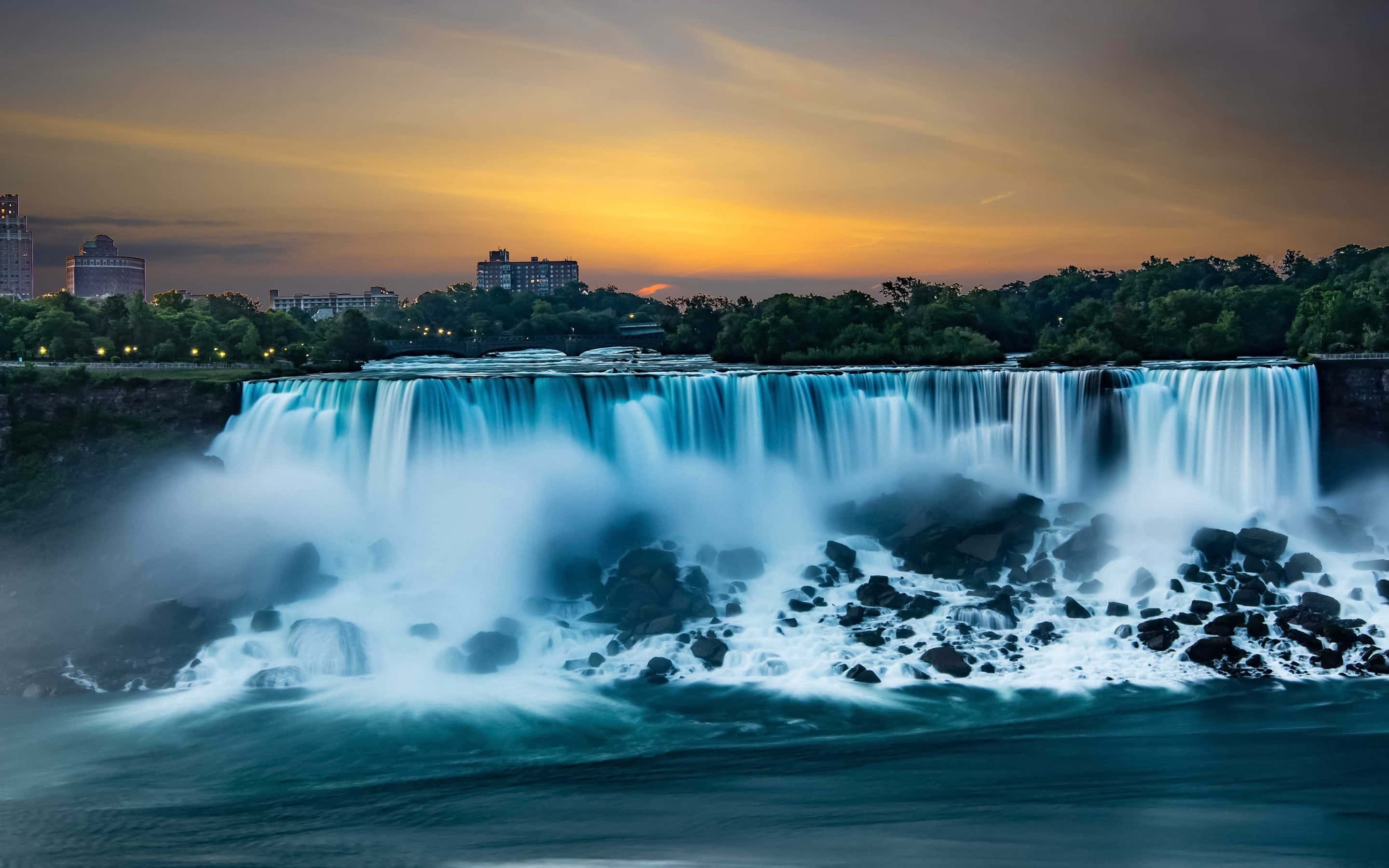Niagara Falls Canada 桌布 壁紙 — 尼亞加拉大瀑布在金色夕陽天空下，以燦爛的藍綠色傾瀉而下，遠方城市燈光閃爍。