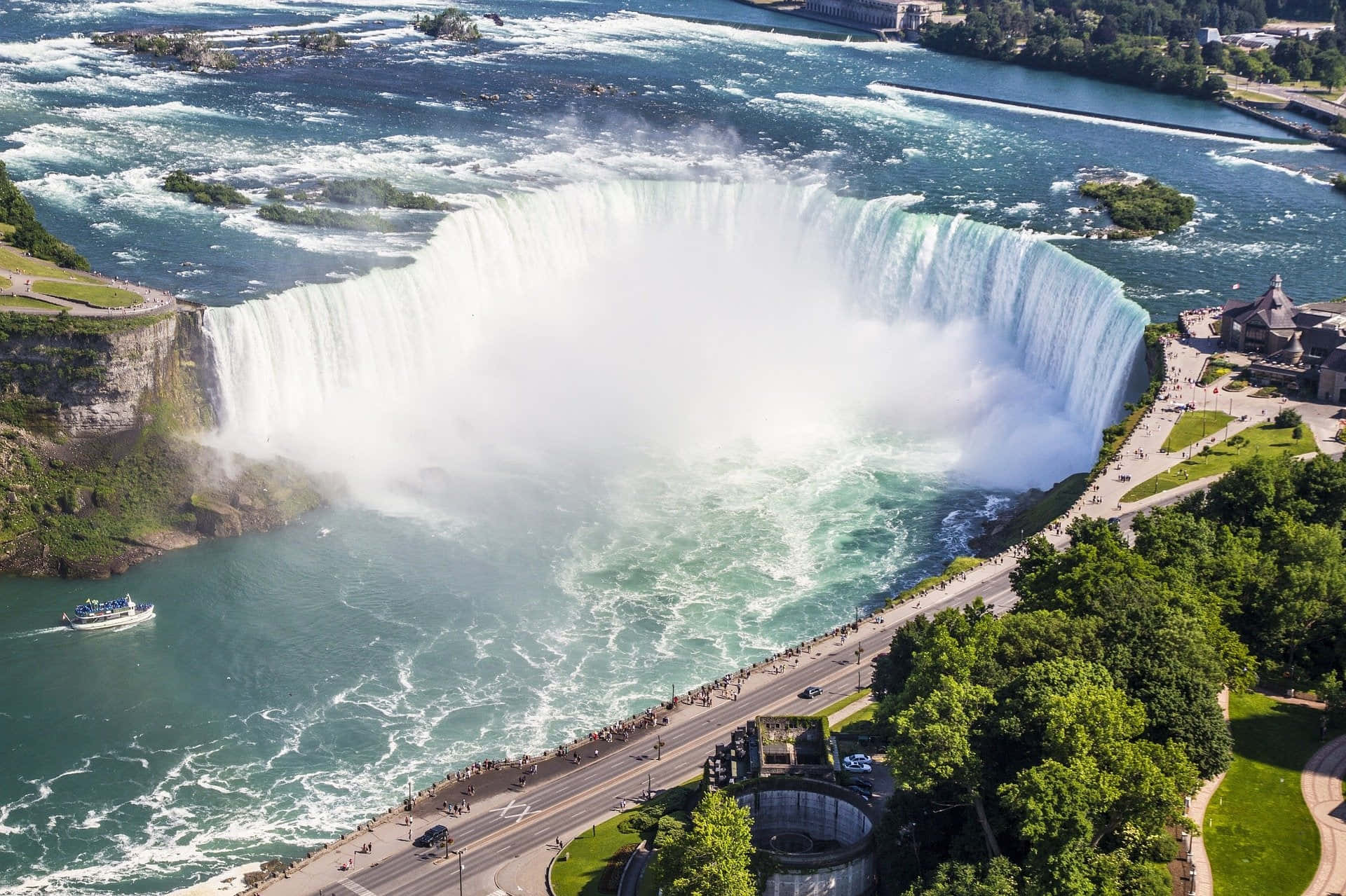 Niagara Falls 桌布 壁紙 — 尼亞加拉大瀑布從空中俯瞰，強勁地傾瀉入一個煙霧繚繞的綠松石色盆地，四周環繞著茂密的綠植和觀光步道。