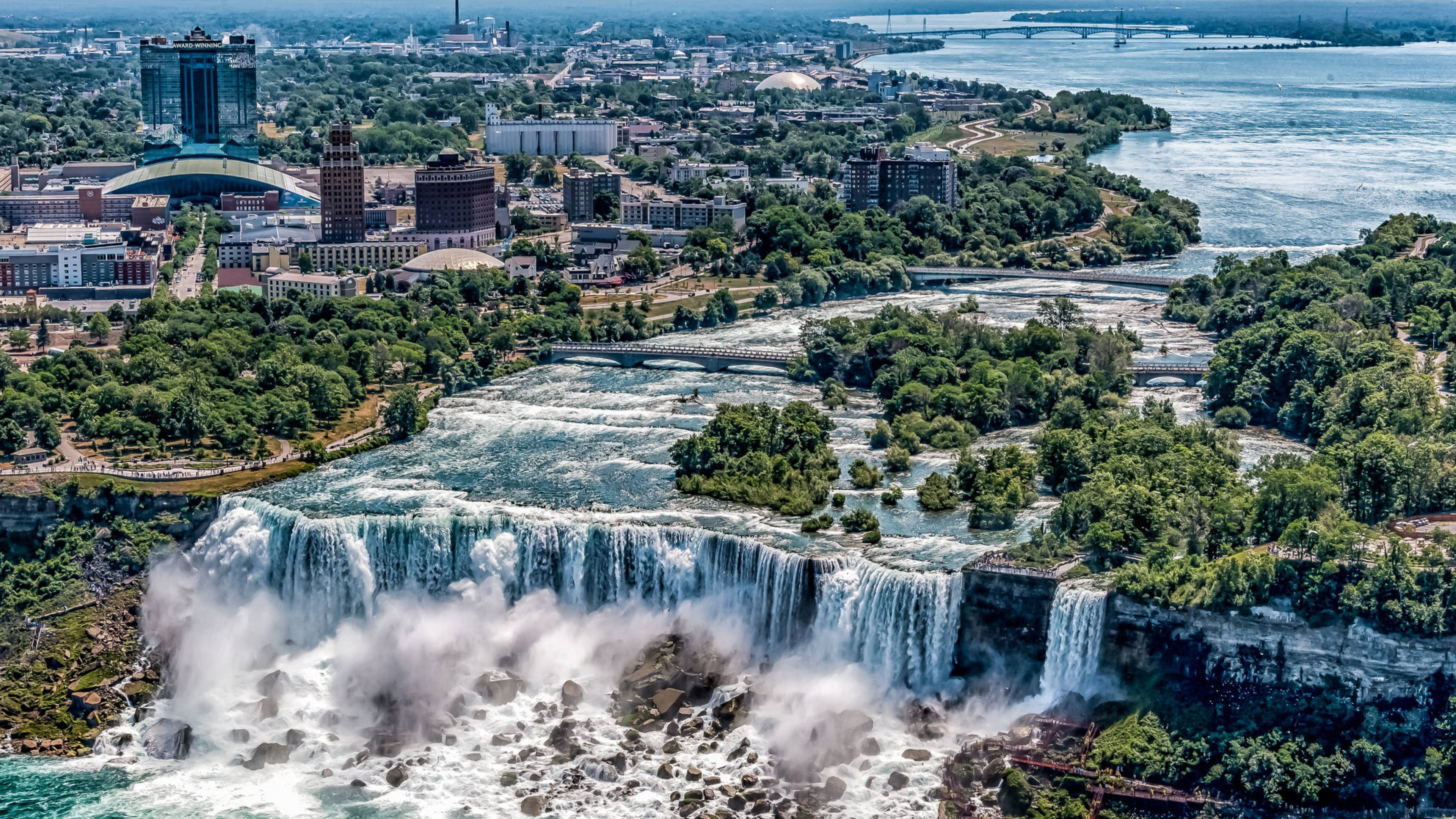 niagara 桌布 壁紙 — 尼亞加拉大瀑布從高空俯瞰，強力傾瀉入霧氣瀰漫的水中，背景是鬱鬱蔥蔥的城市景觀和河流。