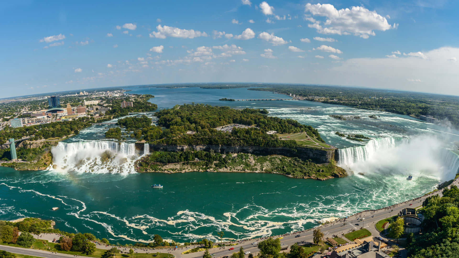 720p Niagara Falls 桌布 壁紙 — 尼亞加拉大瀑布從高空俯瞰，強力傾瀉入碧綠色的水域，上方是明亮的藍天和散落的雲朵。