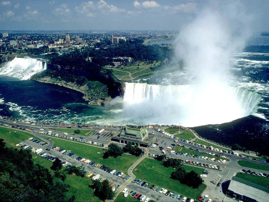 Niagara Falls 桌布 壁紙 — 尼亞加拉大瀑布從高空俯瞰，強勁地傾瀉入霧氣瀰漫的盆地，周圍綠意盎然，下方有停車場。