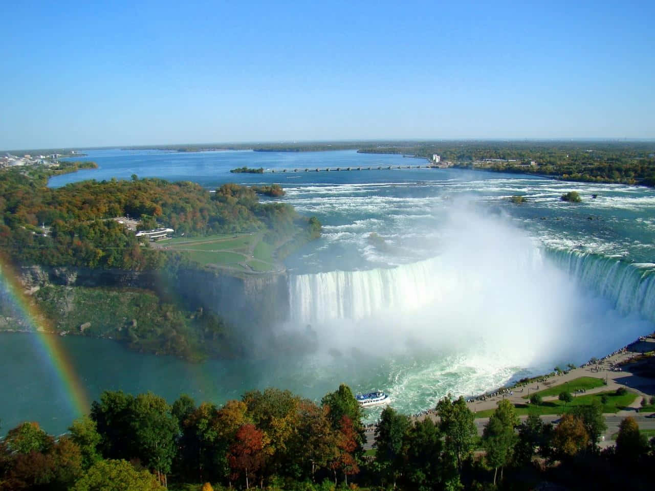 720p Niagara Falls 桌布 壁紙 — 尼亞加拉大瀑布的空中俯瞰，水流強勁傾瀉，霧氣升騰，一道鮮豔的彩虹拱跨在秋色樹木之上。