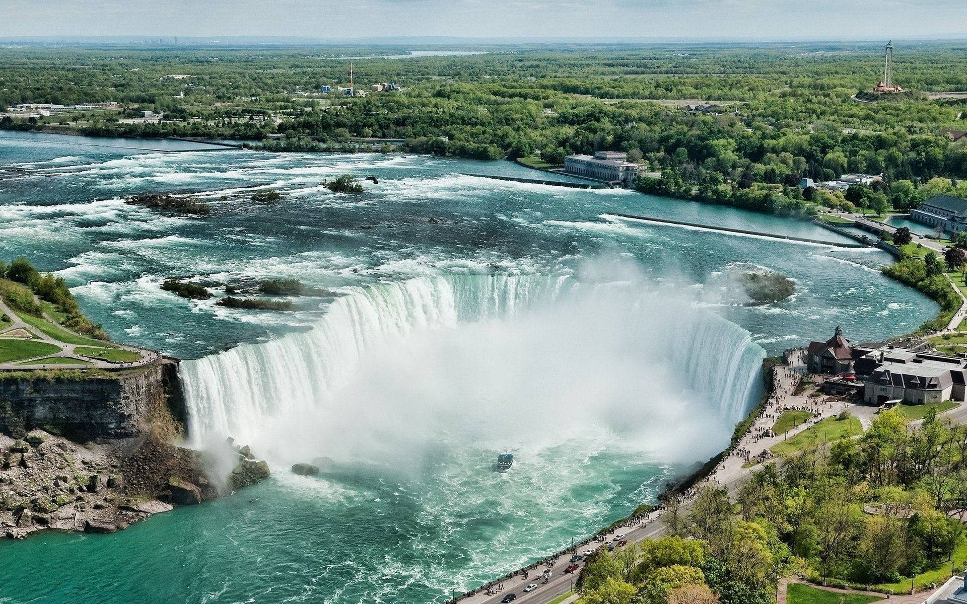 niagara 桌布 壁紙 — 尼亞加拉大瀑布的鳥瞰圖，強力傾瀉入一個霧氣瀰漫的綠松石色盆地，周圍環繞著茂密的綠色森林和旅遊基礎設施。