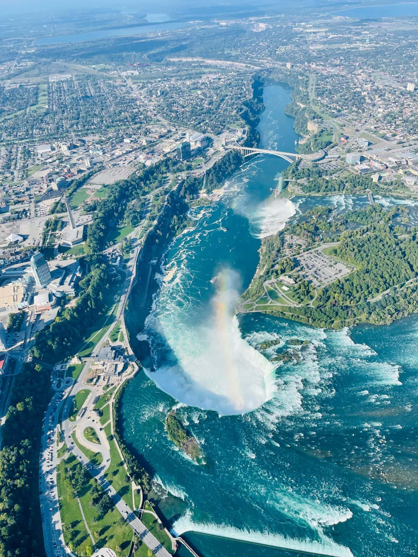Niagara Falls 桌布 壁紙 — 尼亞加拉大瀑布的鳥瞰圖，強勁地傾瀉入藍綠色的河流，一道鮮豔的彩虹在霧氣中拱起，橫跨城市和森林景觀。