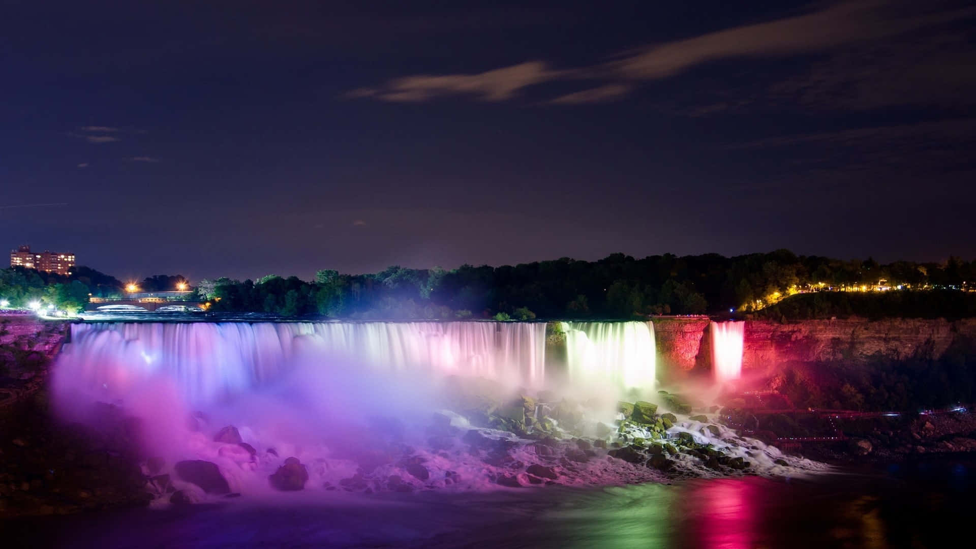 Niagara Falls Canada 桌布 壁紙 — 尼亞加拉瀑布在夜晚被鮮豔的紫色、粉紅色和綠色燈光照亮，在黑暗的天空下反射在霧氣瀰漫的水面上。