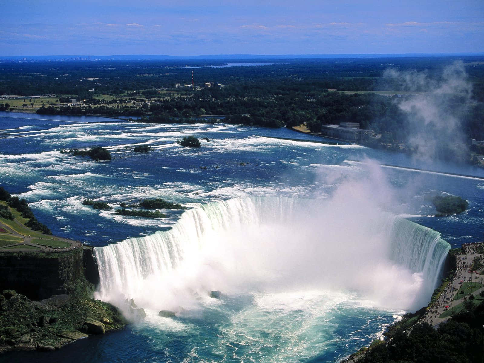 Niagara Falls 桌布 壁紙 — 尼加拉瀑布在晴朗藍天下，從高空俯瞰，強勁地傾瀉入霧氣瀰漫的河流，四周綠意盎然，遠方城市景觀依稀可見。
