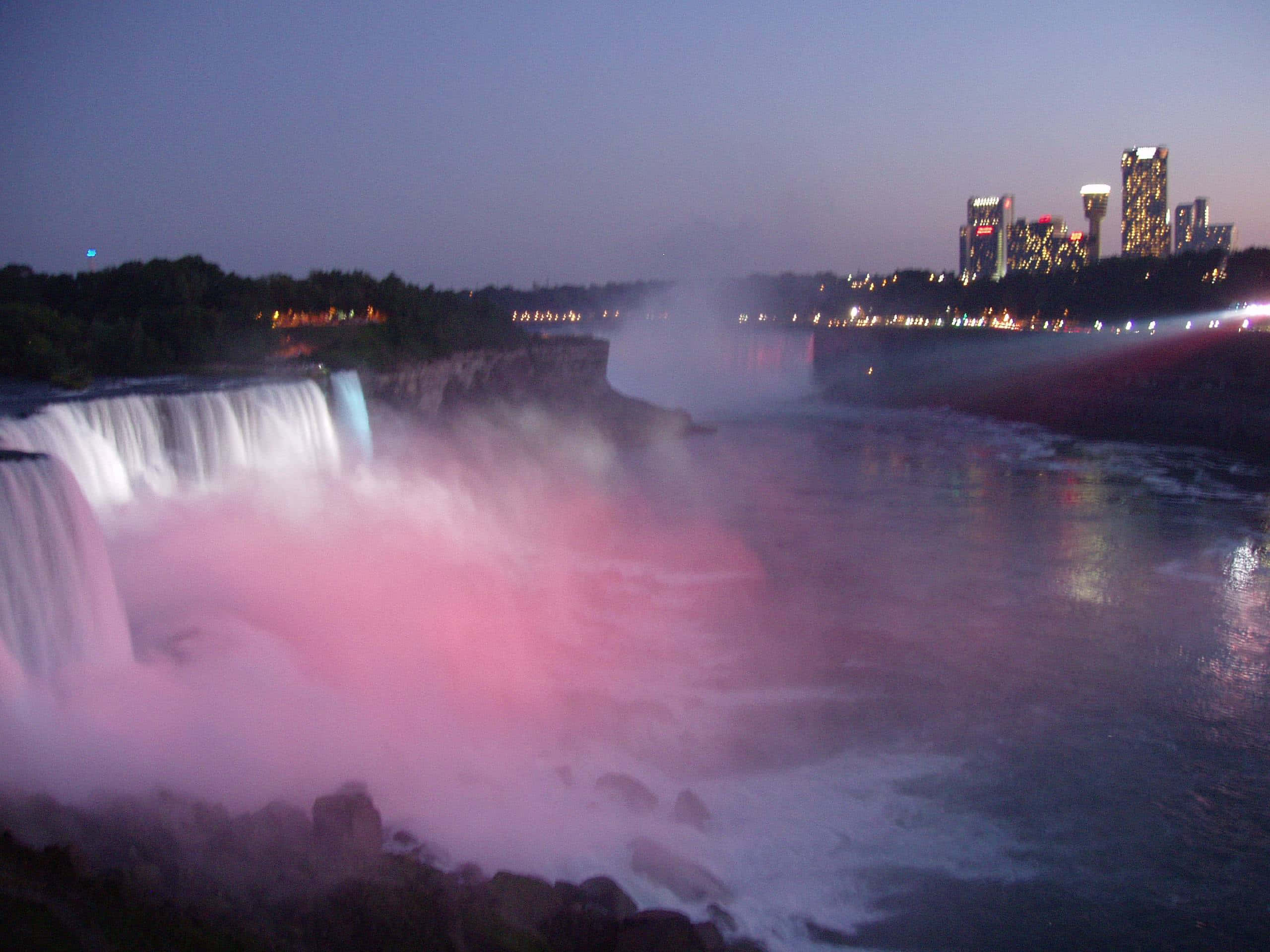 Niagara Falls 桌布 壁紙 — 尼加拉瀑布在黃昏時被粉紅燈光照亮，背景中城市天際線閃爍著光芒。