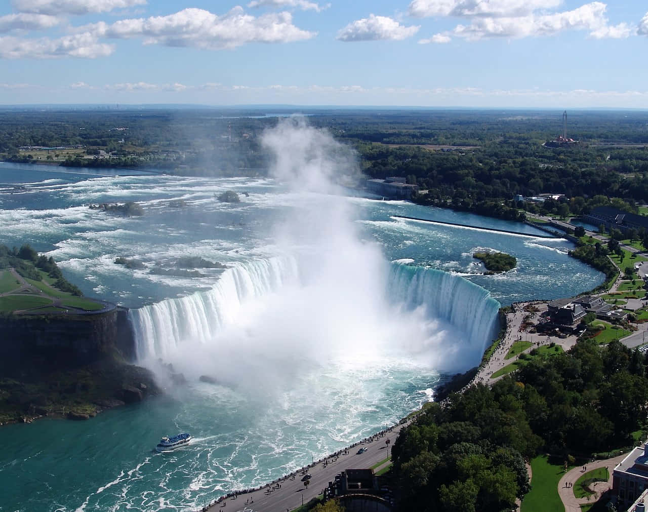 Niagara Falls 桌布 壁紙 — 尼加拉瀑布的鳥瞰圖，在明亮的藍天下，瀑布強勁地傾瀉入一個霧氣瀰漫的盆地，周圍環繞著茂密的綠植和旅遊步道。