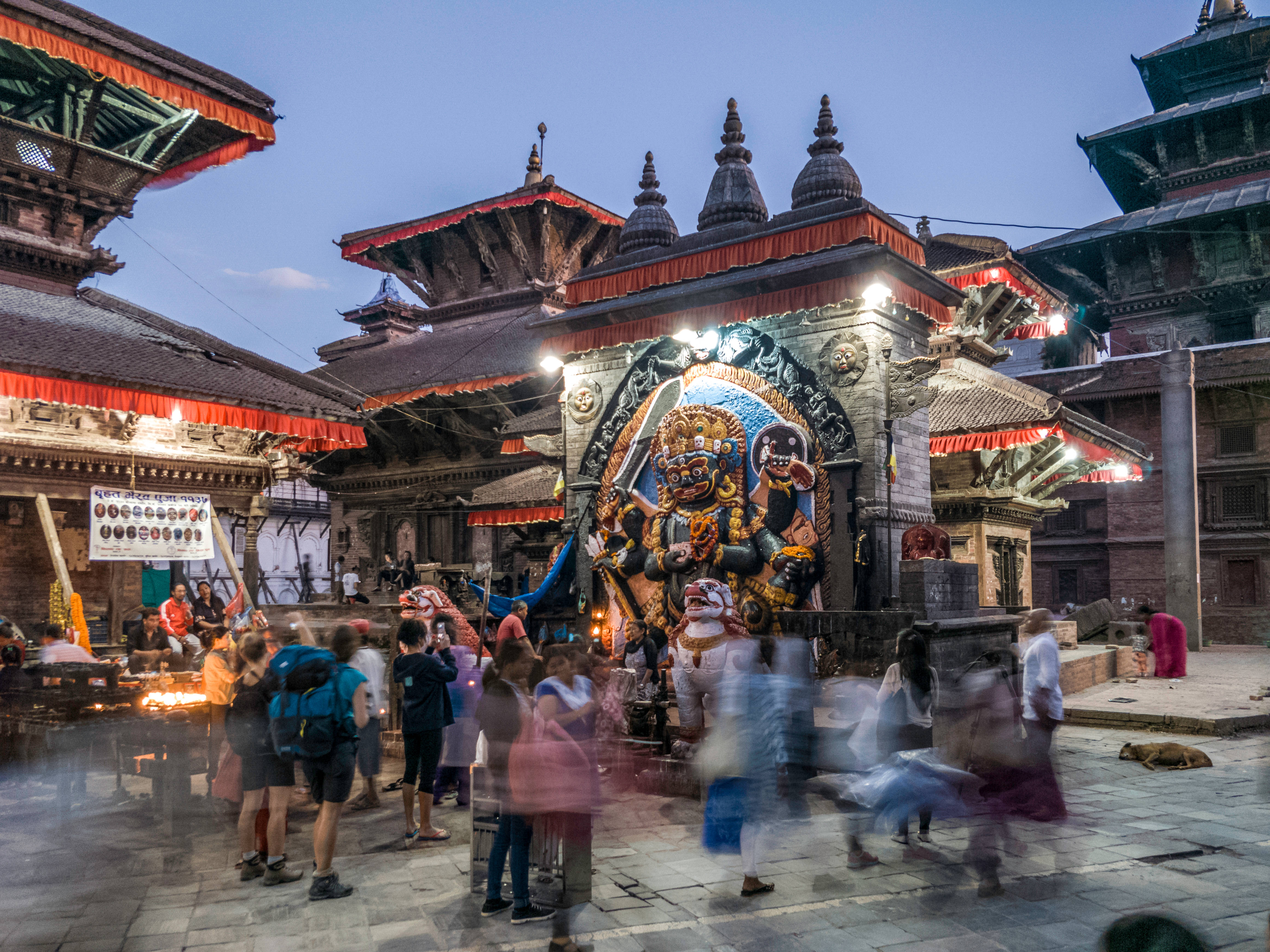 Nepal 桌布 壁紙 — 尼泊爾寺廟廣場上充滿活力的黃昏場景，有一座大型神像和模糊的信眾走動。