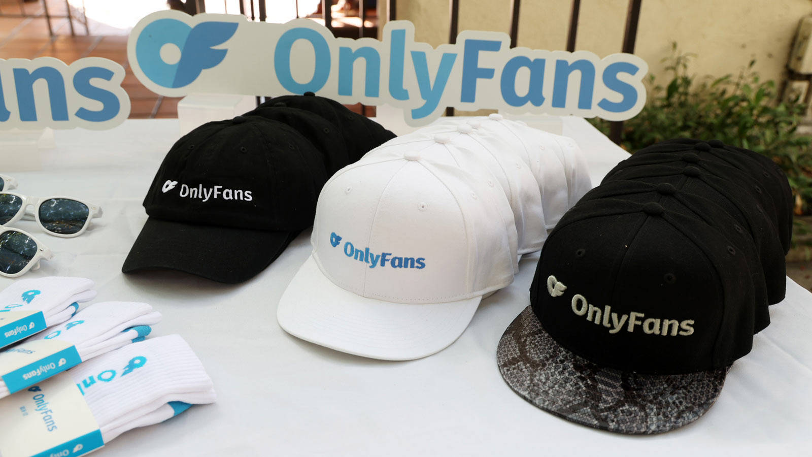 品牌 桌布 壁紙 — 展示OnlyFans品牌商品，包括黑白帽子和襪子，擺放在白色桌子上，背景有一個大型OnlyFans標誌。
