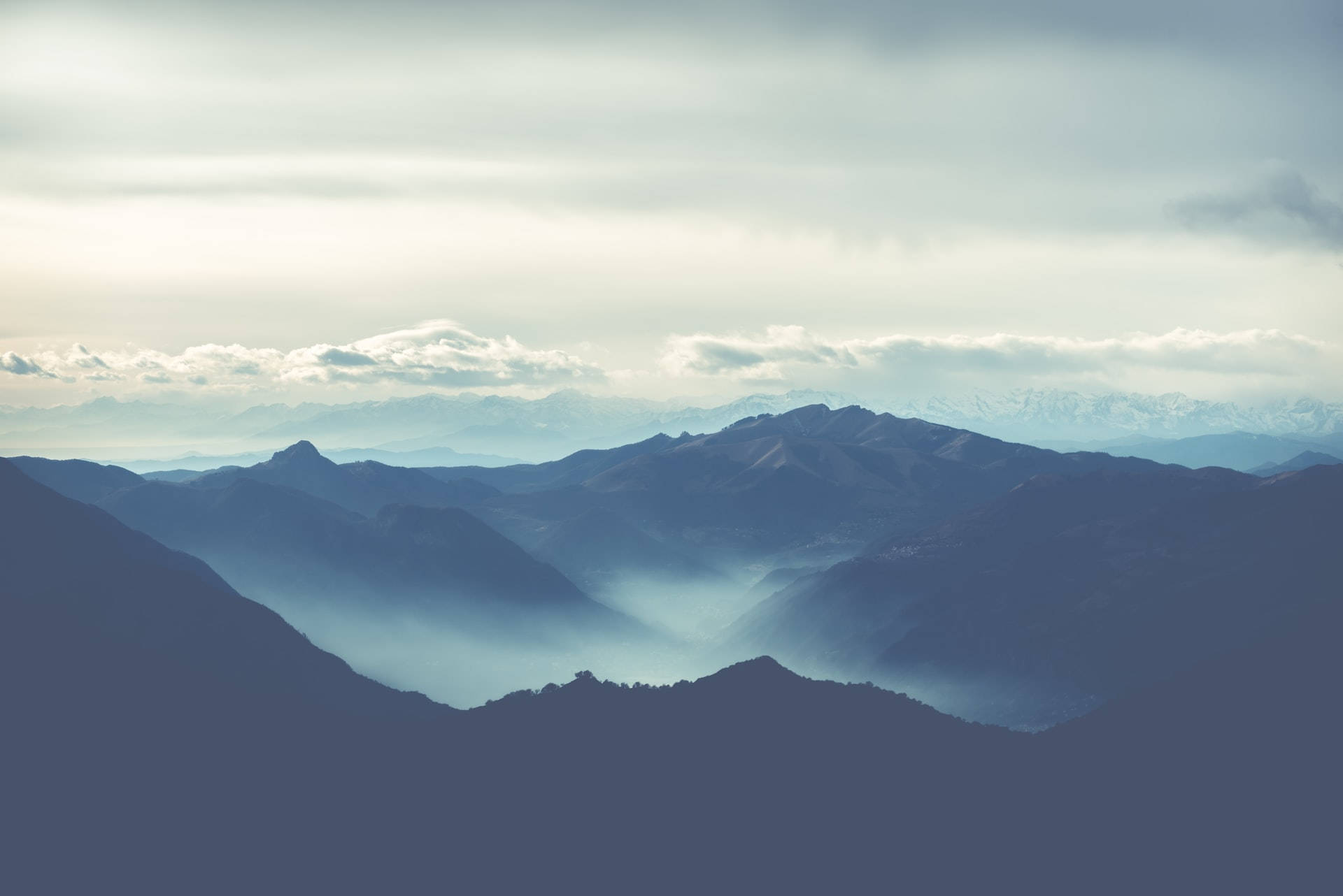 Mountain Landscape 桌布 壁紙 — 層層疊疊的山脈在柔和的多雲天空下逐漸消失在霧氣中，遠處有積雪的山峰。