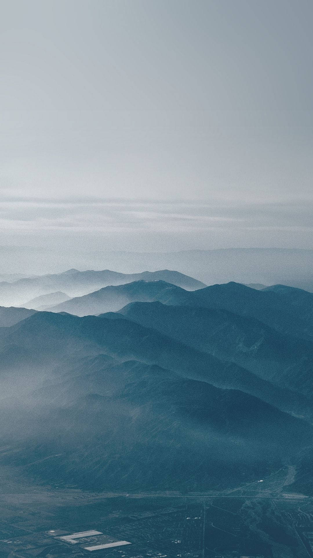 View 桌布 壁紙 — 層層疊疊的山脈在蒼白的天空下逐漸消失在霧氣中，下方是遙遠的山谷。