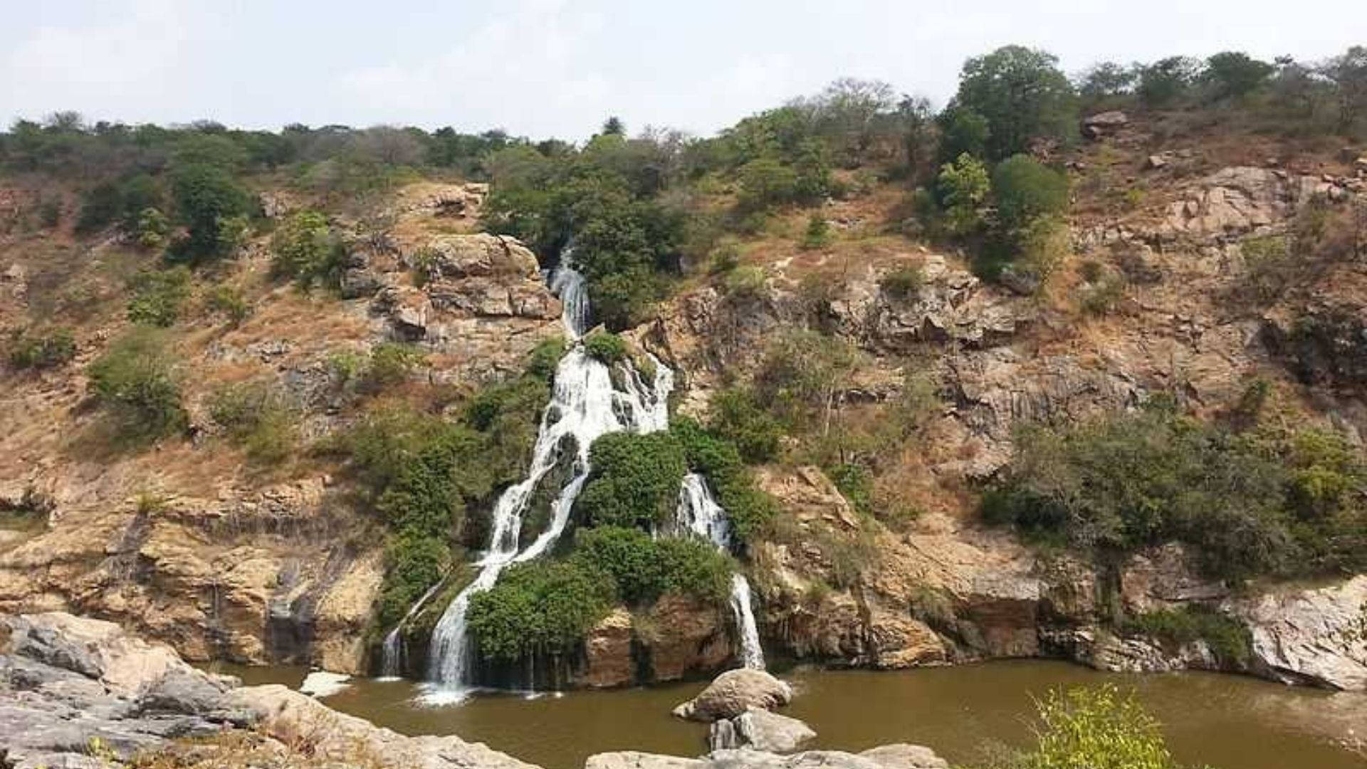 Bangalore 桌布 壁紙 — 層疊瀑布從岩石懸崖傾瀉而下，流入被乾燥植被環繞的平靜水池，天空多雲。