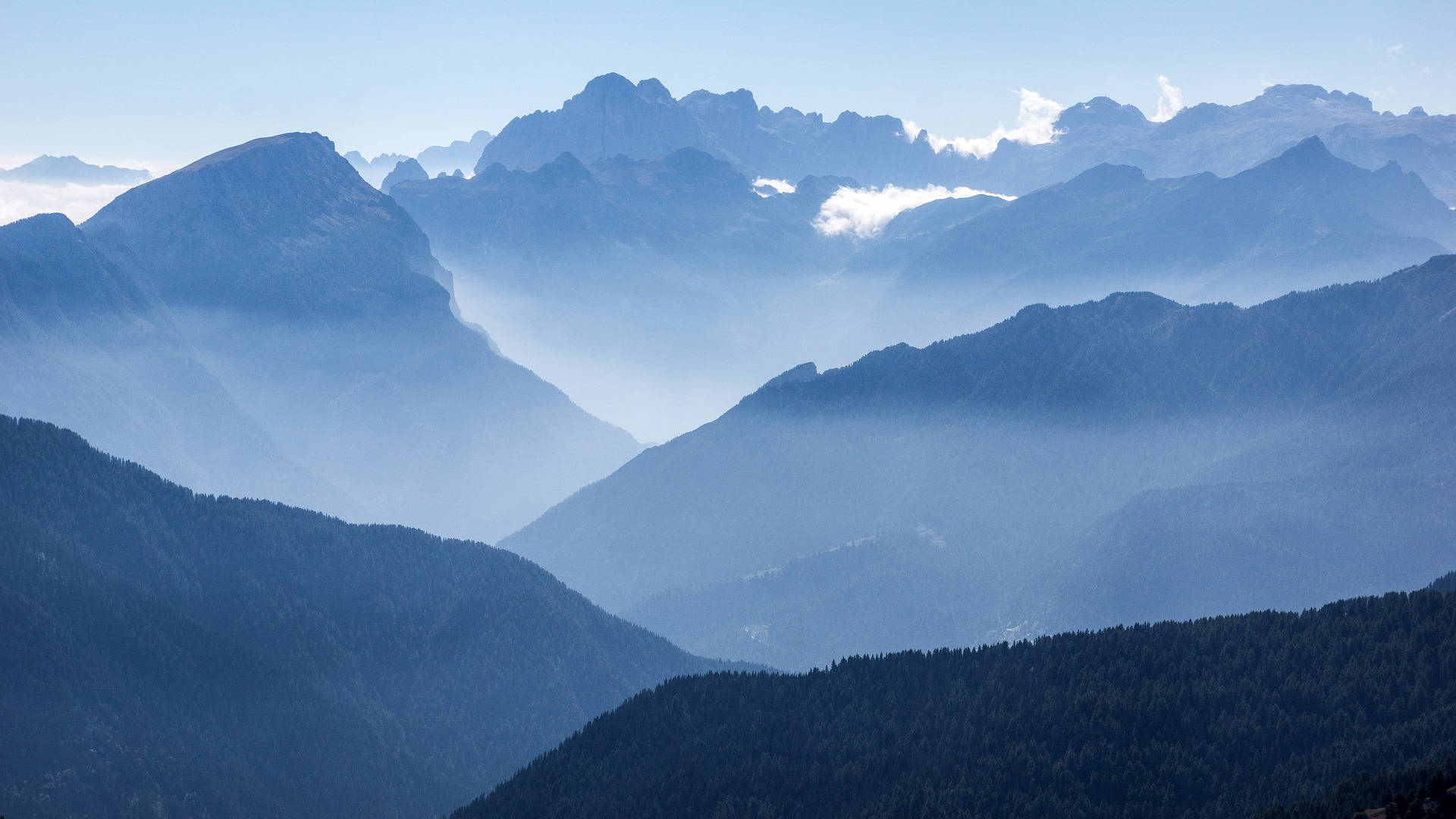 Beautiful Mountain View 桌布 壁紙 — 層疊的山峰在晴朗的藍天下逐漸消失在霧氣中，前景是森林覆蓋的山坡。