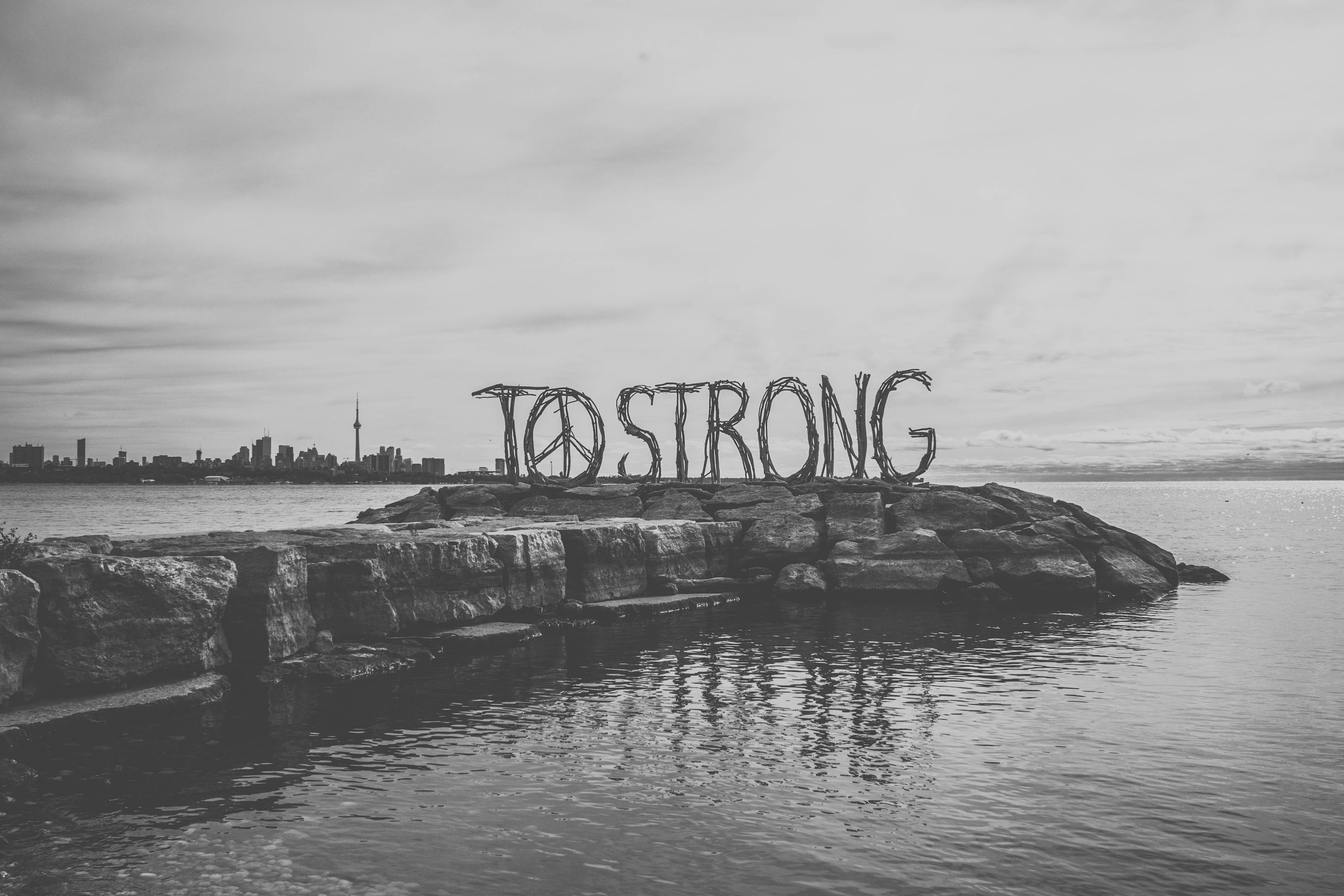 和平符號 桌布 壁紙 — 岩石島上「TO STRONG」編織標誌的黑白景觀，多倫多天際線倒映在平靜的水面上。