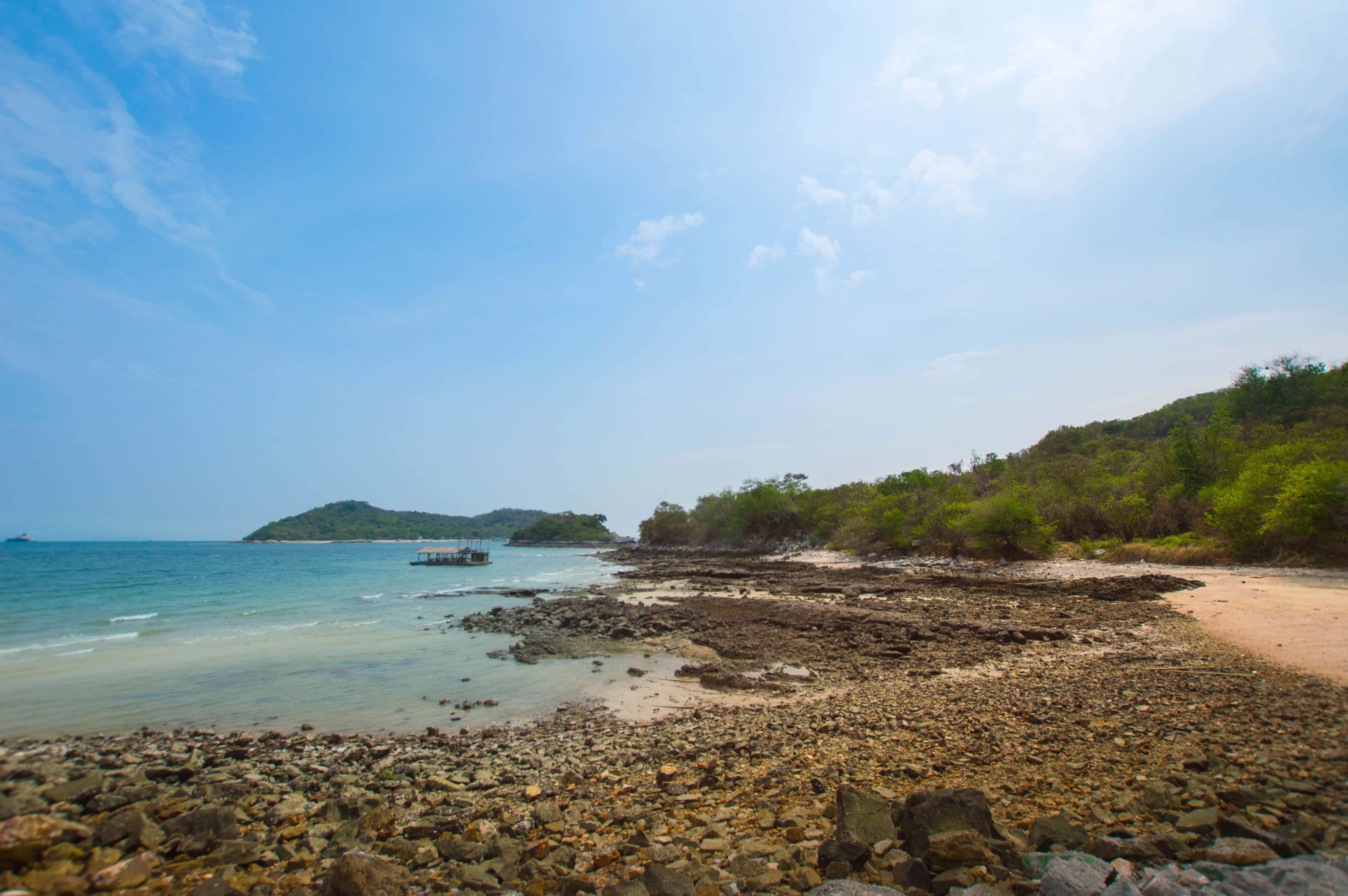 Beach Sand 桌布 壁紙 — 岩石熱帶海岸線，有碧綠海水、柔和波浪和茂密綠色山丘，在明亮的藍天下。