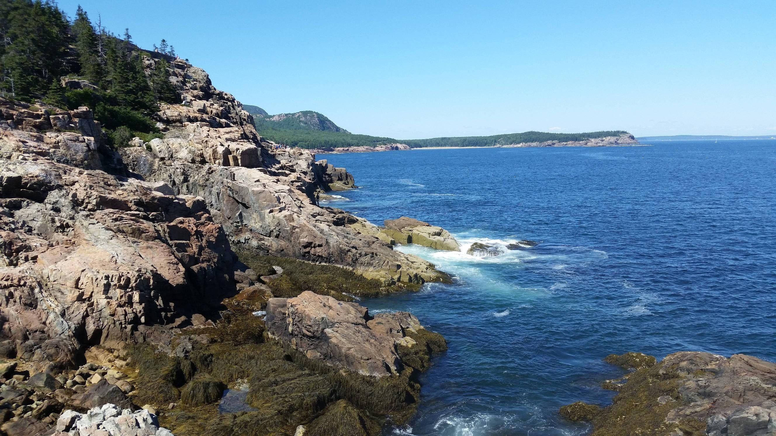 自然 桌布 壁紙 — 崎嶇岩石海岸線，在清澈藍天下有拍岸浪濤，遠方是森林覆蓋的山丘。