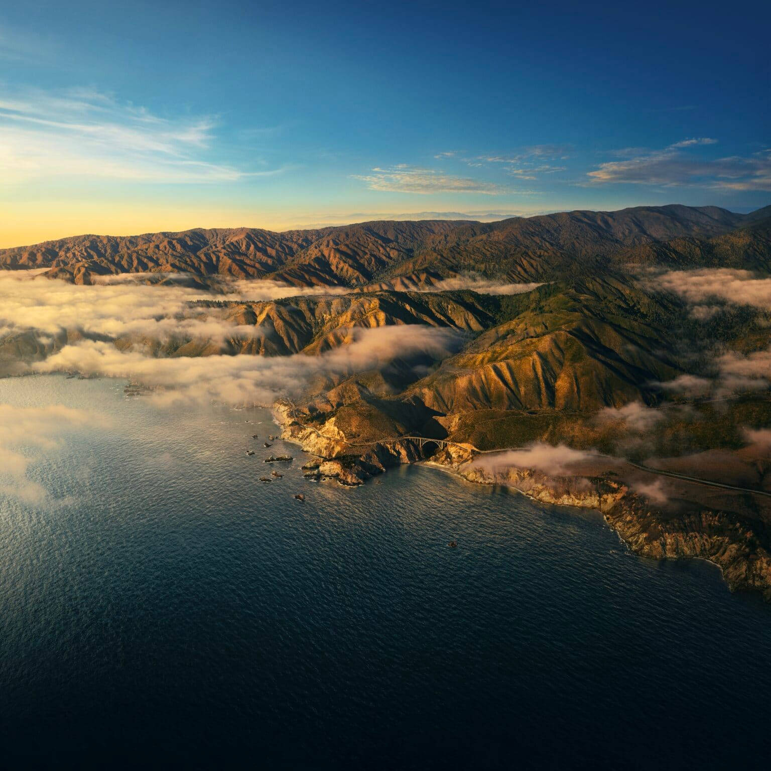 山景 桌布 壁紙 — 崎嶇海岸山脈的鳥瞰圖，沐浴在金色日出光芒中，雲霧繚繞山谷，下方是深藍色海洋。