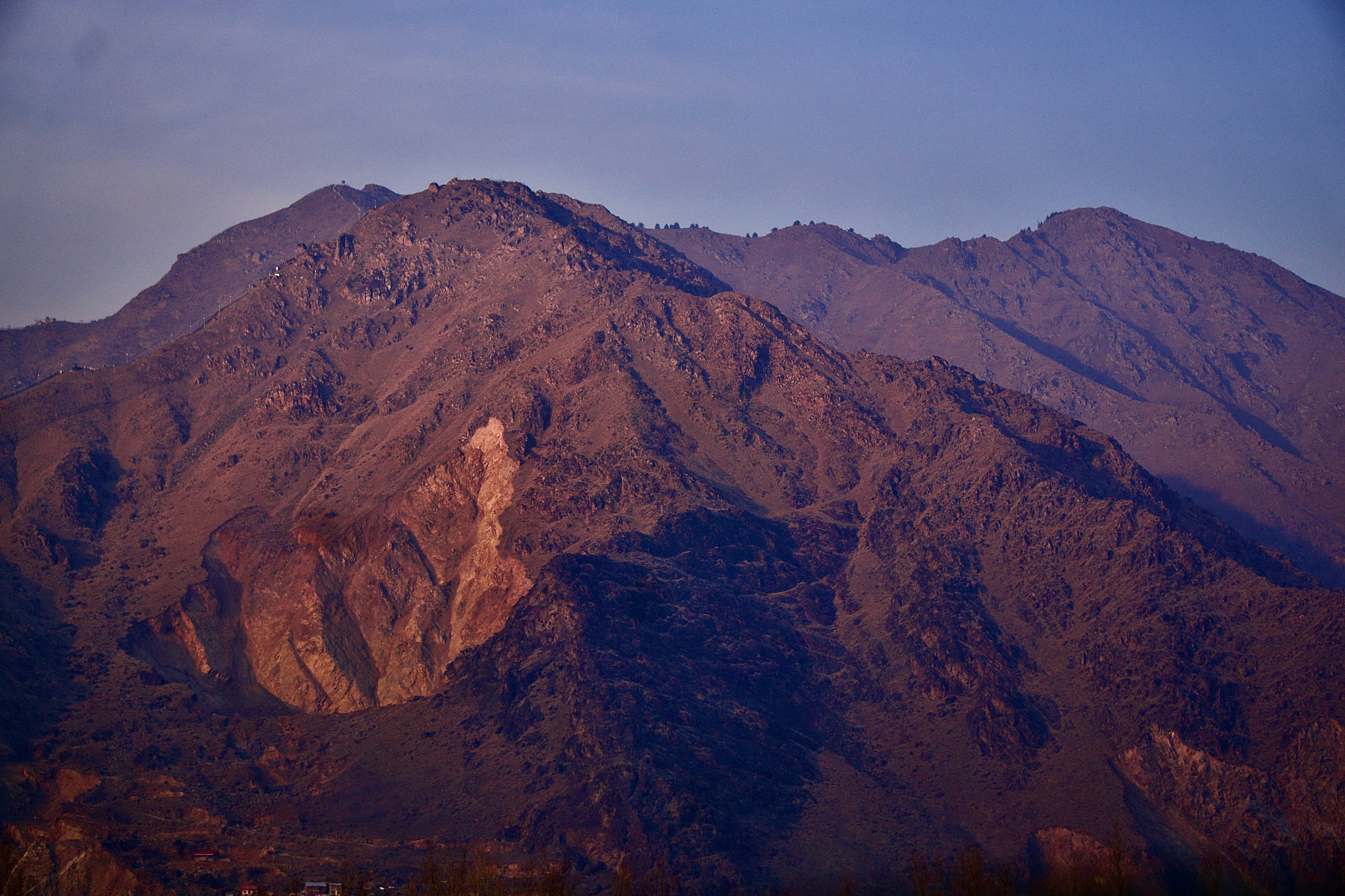 Topography 桌布 壁紙 — 崎嶇的山峰沐浴在溫暖的日落光芒中，在柔和的藍天下，光禿的山坡上投下長長的影子。