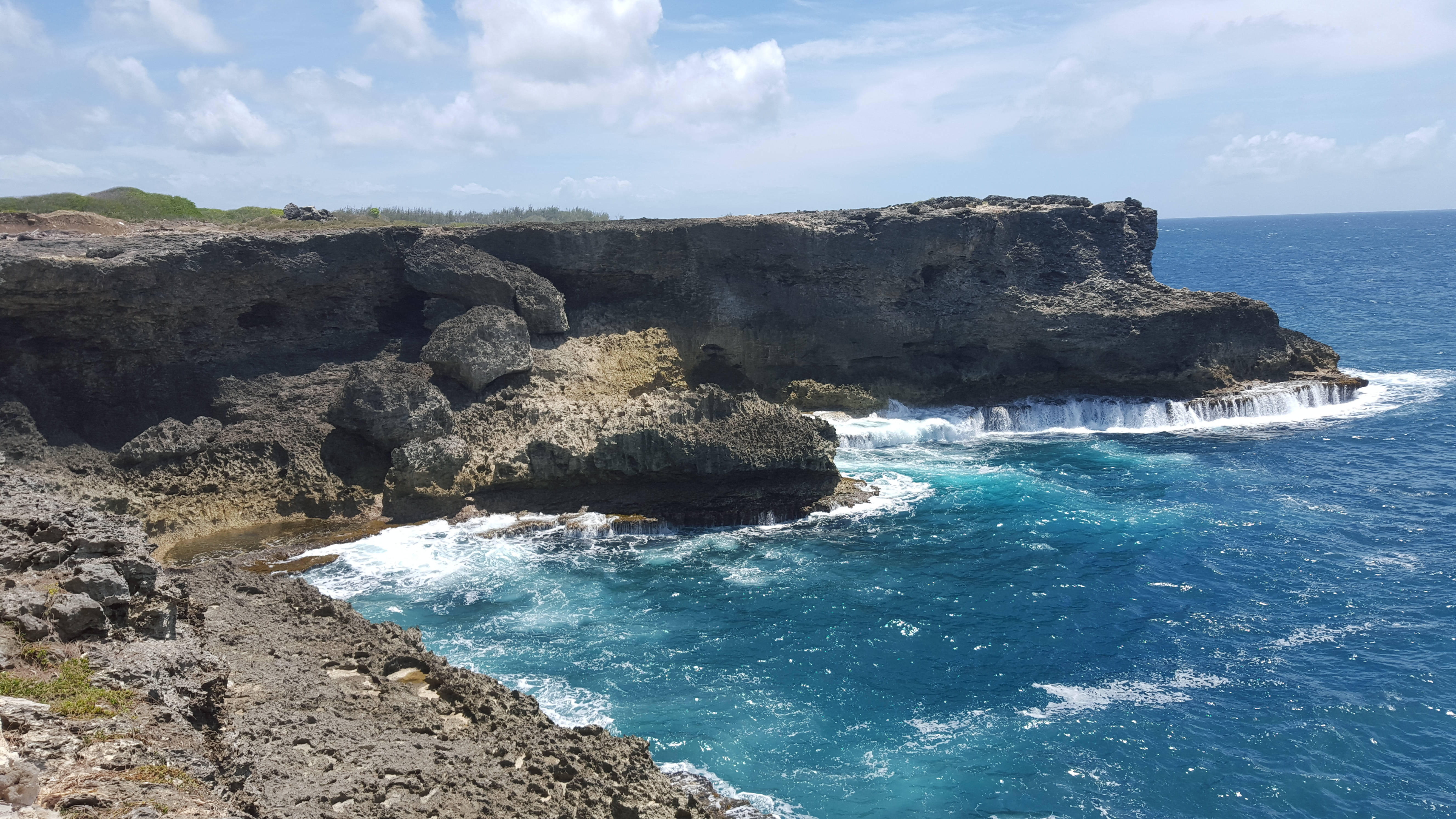 Barbados 桌布 壁紙 — 崎嶇的懸崖陡峭地插入碧綠的海水中，海浪在明亮的藍天下拍打著岩石海岸。
