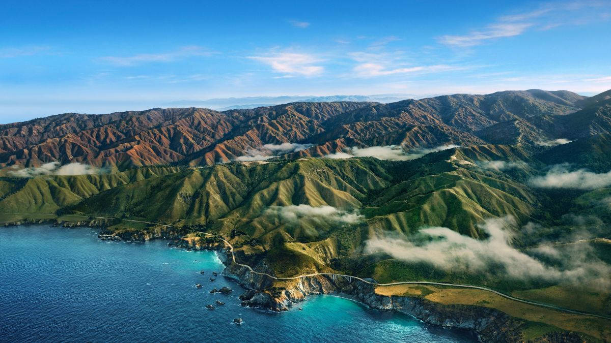 山景 桌布 壁紙 — 崎嶇的綠色山脈與碧綠海洋交匯的鳥瞰圖，蜿蜒的海岸道路和清澈藍天下飄浮的雲朵。