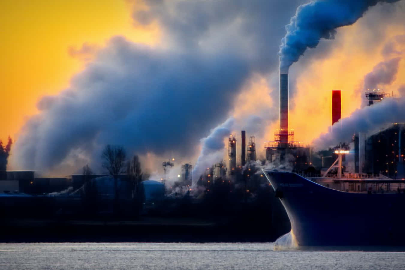 Pollution 桌布 壁紙 — 工業煙囪在熾熱的日落天空下噴出濃煙，一艘船在下方平靜的水面上滑行。