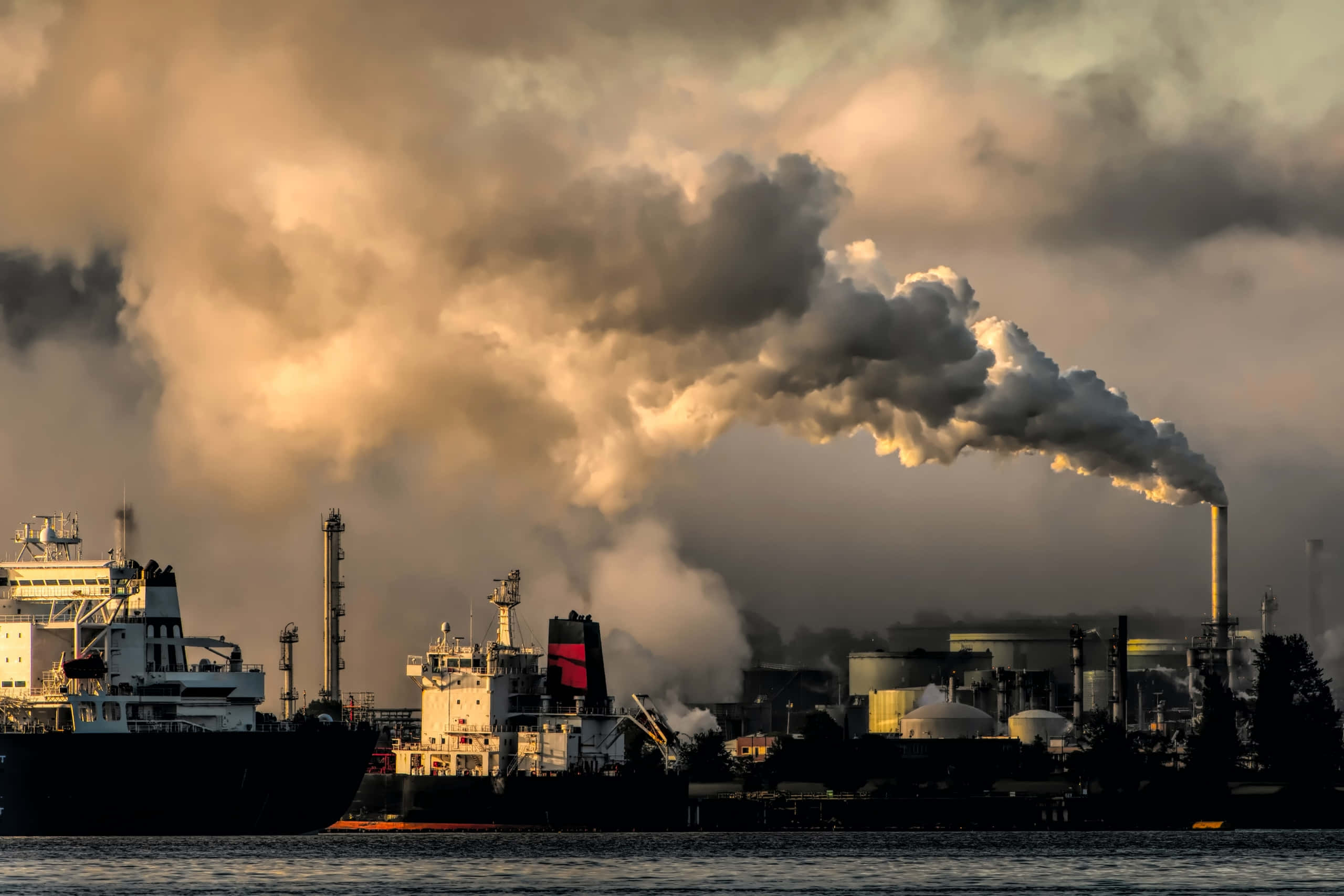 Polluting 桌布 壁紙 — 工業煙霧從工廠煙囪冒出，籠罩著黃金時段的海濱，在船隻和儲油罐上投下戲劇性的陰影。