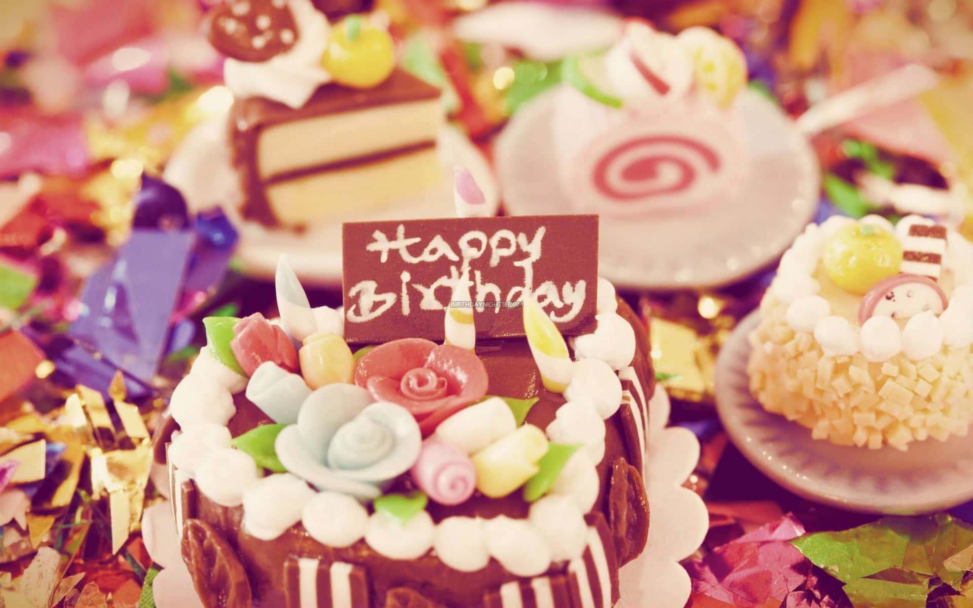 Birthday 桌布 壁紙 — 巧克力生日蛋糕的特寫，裝飾著色彩繽紛的糖花和「Happy Birthday」標牌，周圍環繞著節日五彩紙屑和其他甜點。