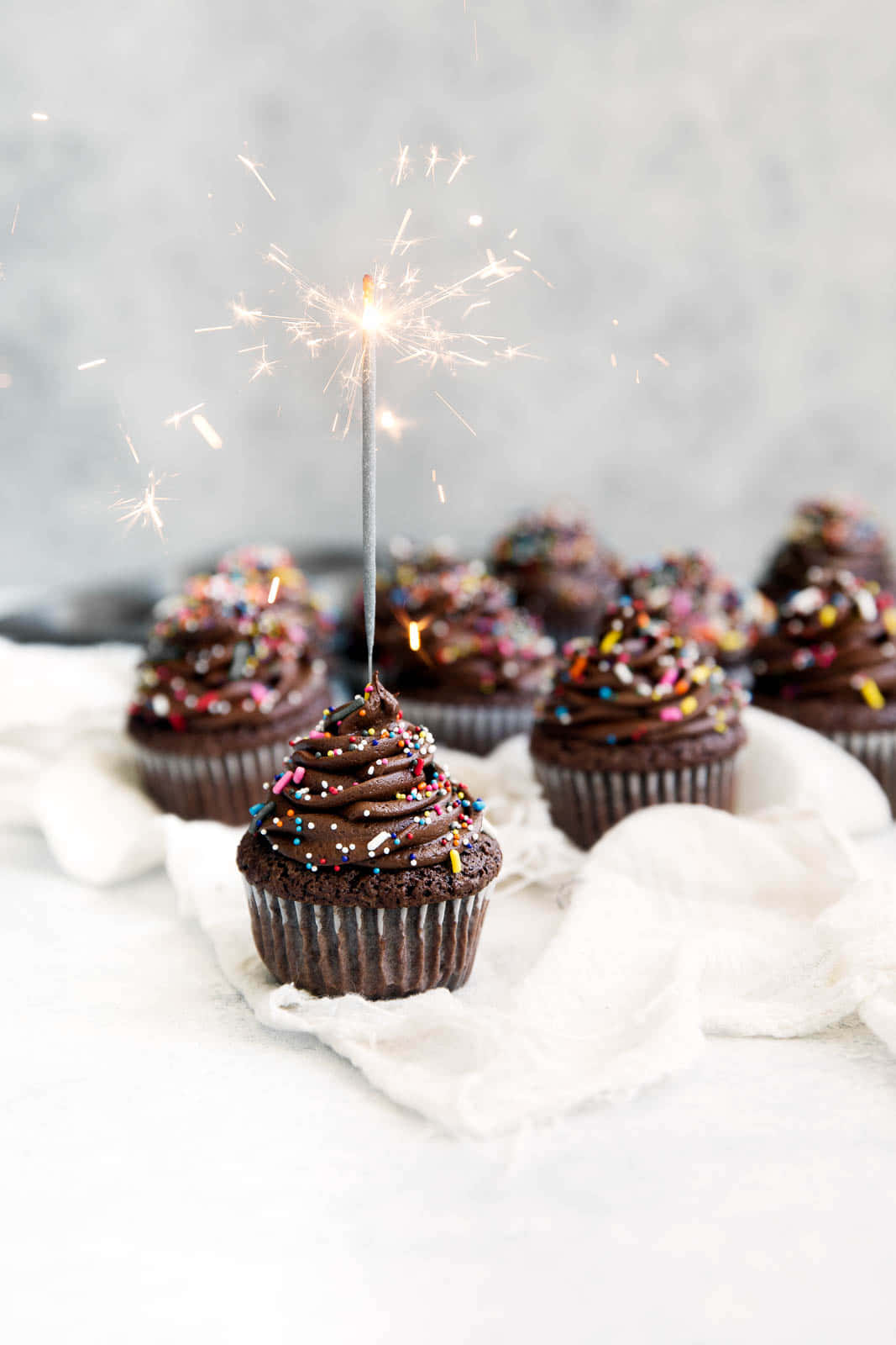 Cupcake 桌布 壁紙 — 巧克力紙杯蛋糕，上面有彩色糖粒和點燃的煙火棒，放在白色布上，背景是柔和的灰色。