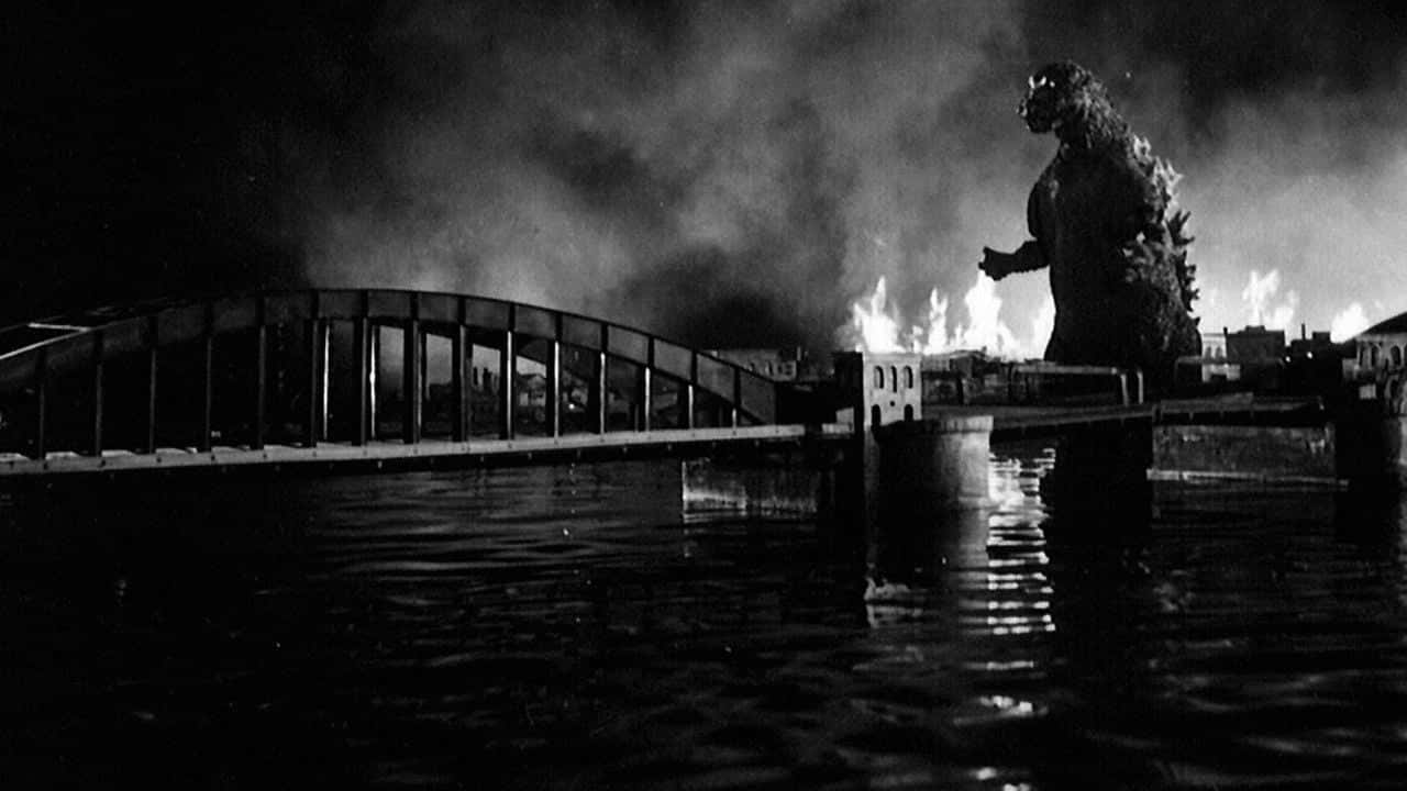 Godzilla 1954 桌布 壁紙 — 巨型怪獸哥吉拉夜晚站在燃燒的橋上，火焰和煙霧在淹沒的城市景觀上升起。