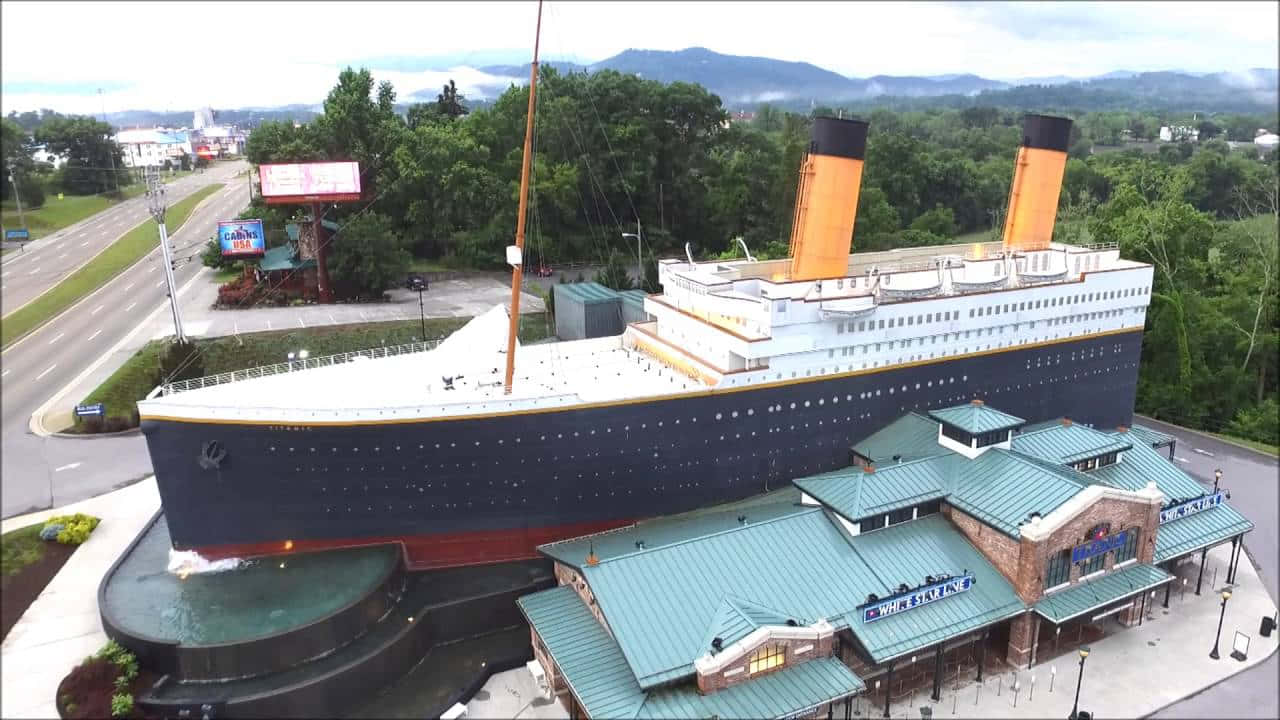 Titanic 桌布 壁紙 — 巨型鐵達尼號複製建築物的鳥瞰圖，設有橙色煙囪和藍色船體，周圍環繞著綠樹和一條道路。
