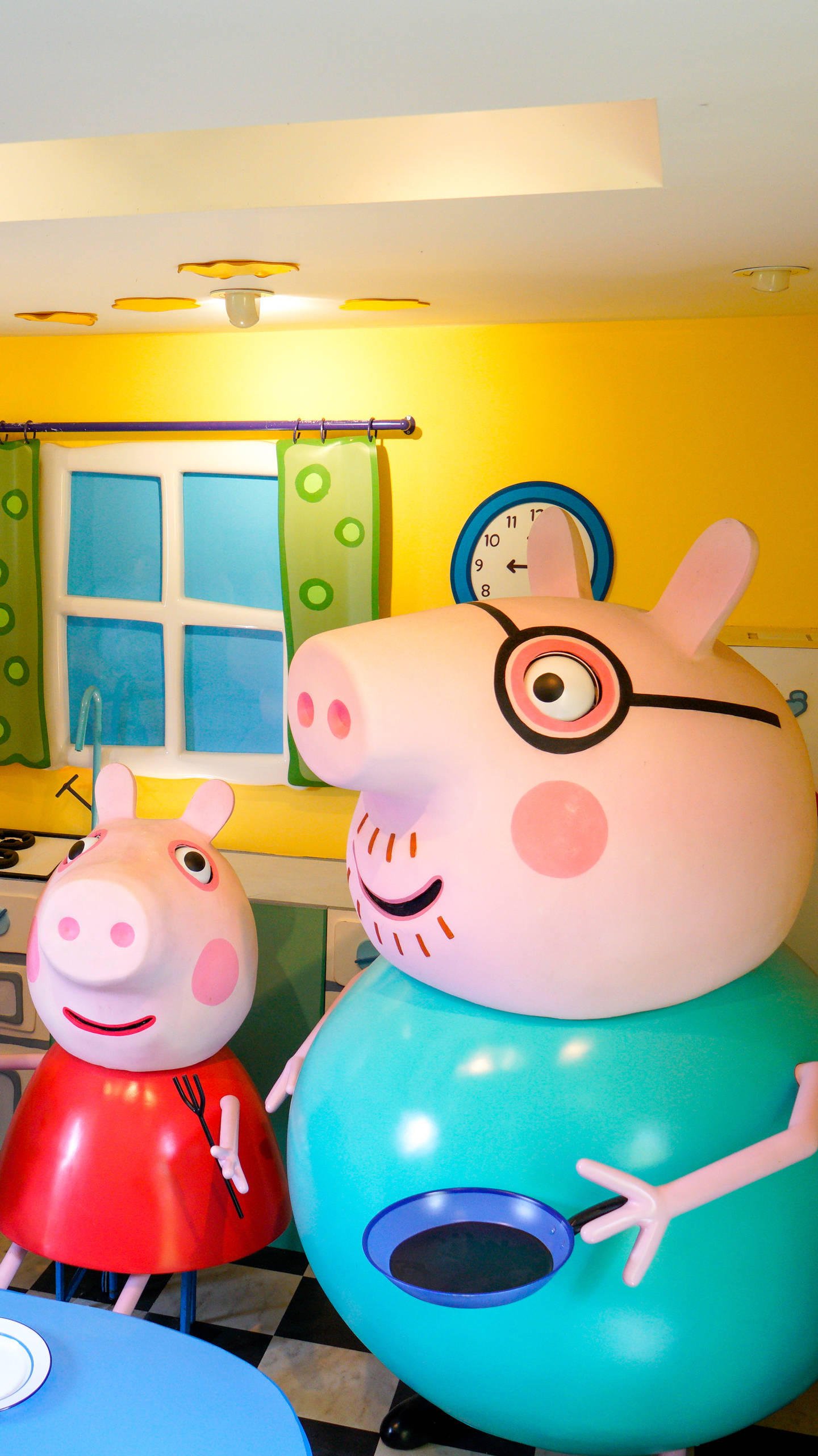Peppa 桌布 壁紙 — 巨大的Peppa Pig和Daddy Pig雕像在一個光線明亮的黃色廚房裡，有格子地板和窗戶。