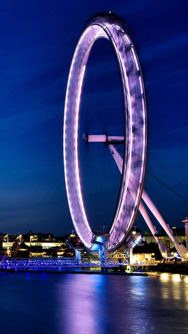 England 桌布 壁紙 — 巨大的倫敦眼在深藍色夜空下發出紫色光芒，燈光倒映在下方平靜的水面上。