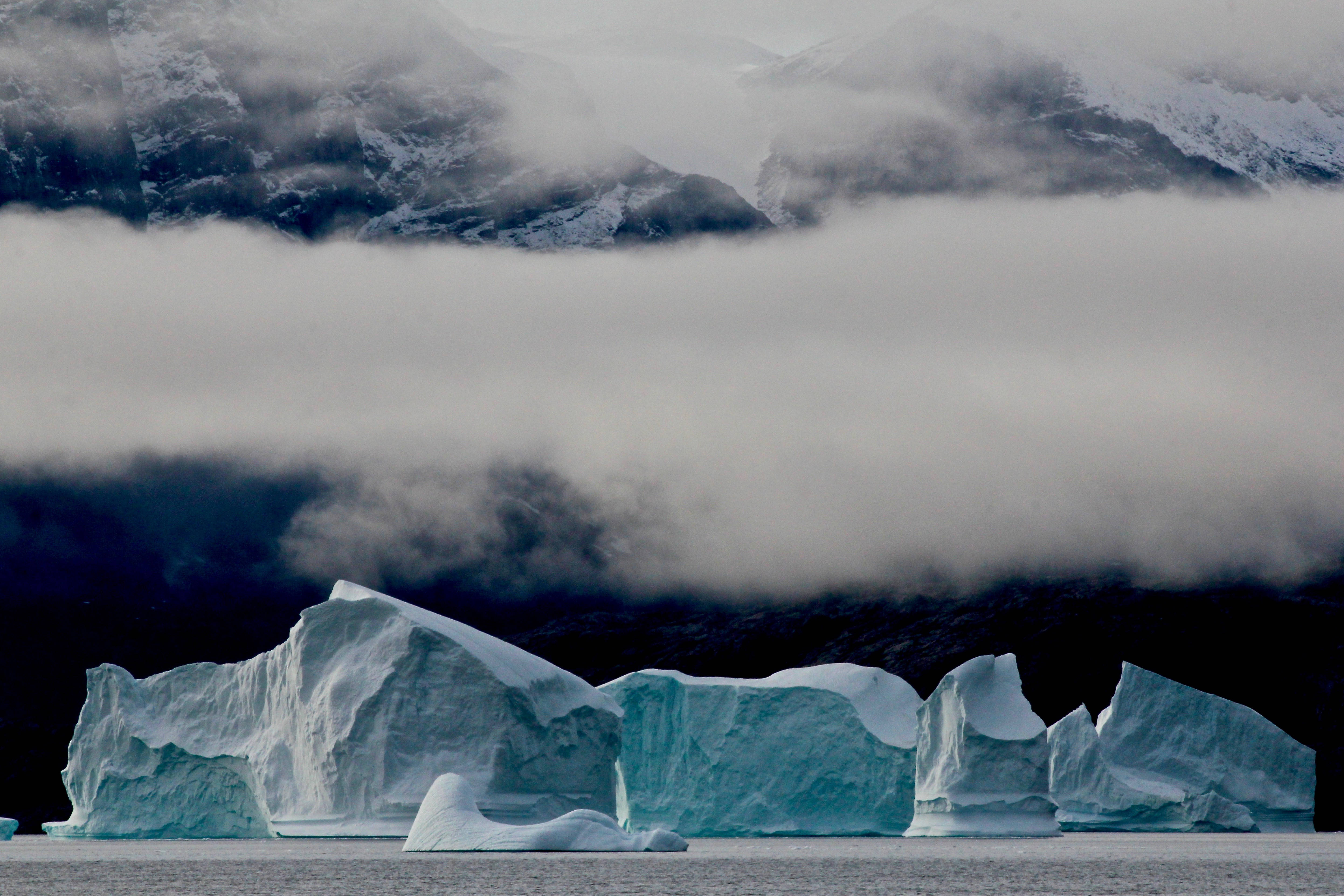 Greenland 桌布 壁紙 — 巨大的冰山帶有藍色內部，在厚厚霧雲下的平靜水域中靜靜漂浮。