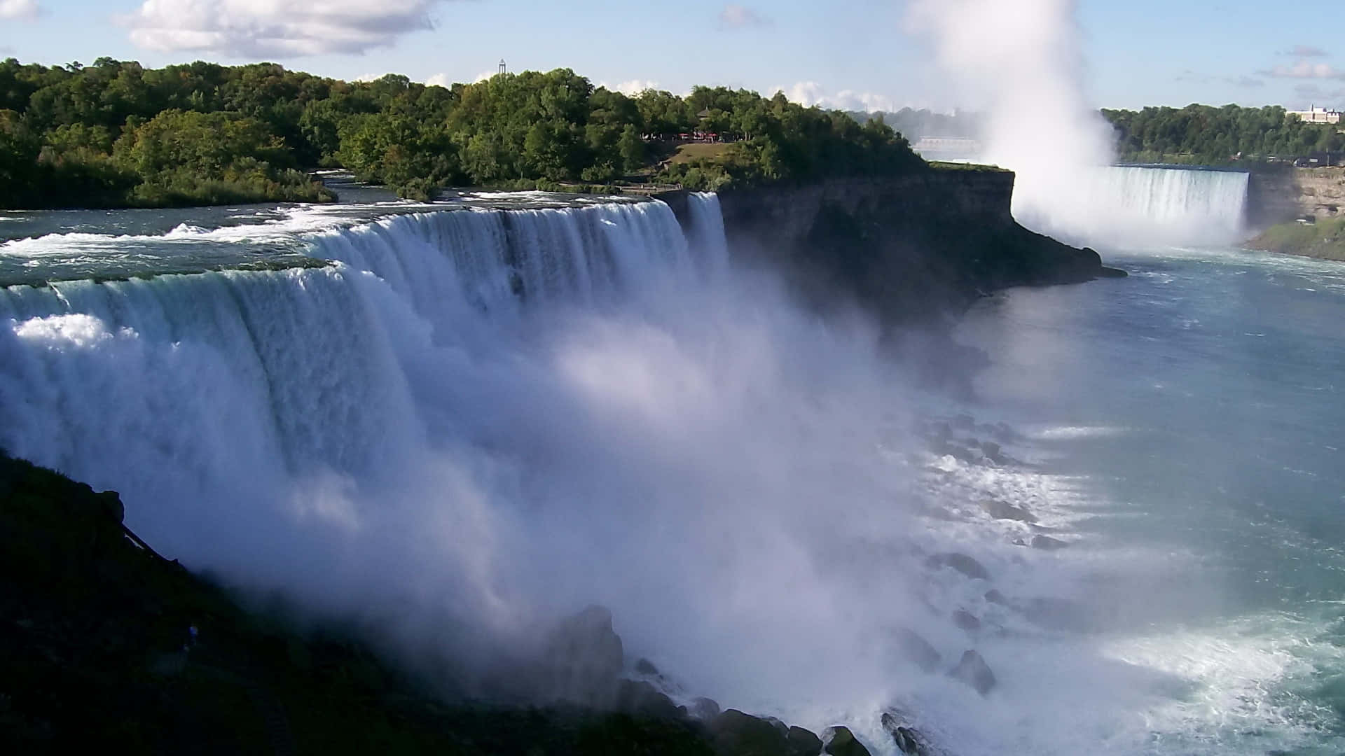 720p Niagara Falls 桌布 壁紙 — 巨大的層疊瀑布從懸崖上咆哮著落入霧氣瀰漫的河流，周圍是茂密的綠色森林，天空明亮。