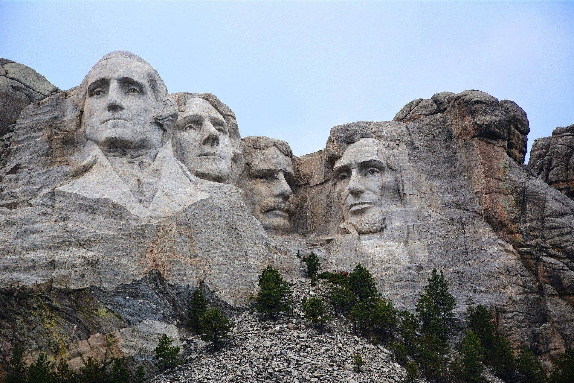 Mount Rushmore 桌布 壁紙 — 巨大的山紀念碑，在晴朗的天空下，岩石上雕刻著四位總統的面孔，下方有松樹。