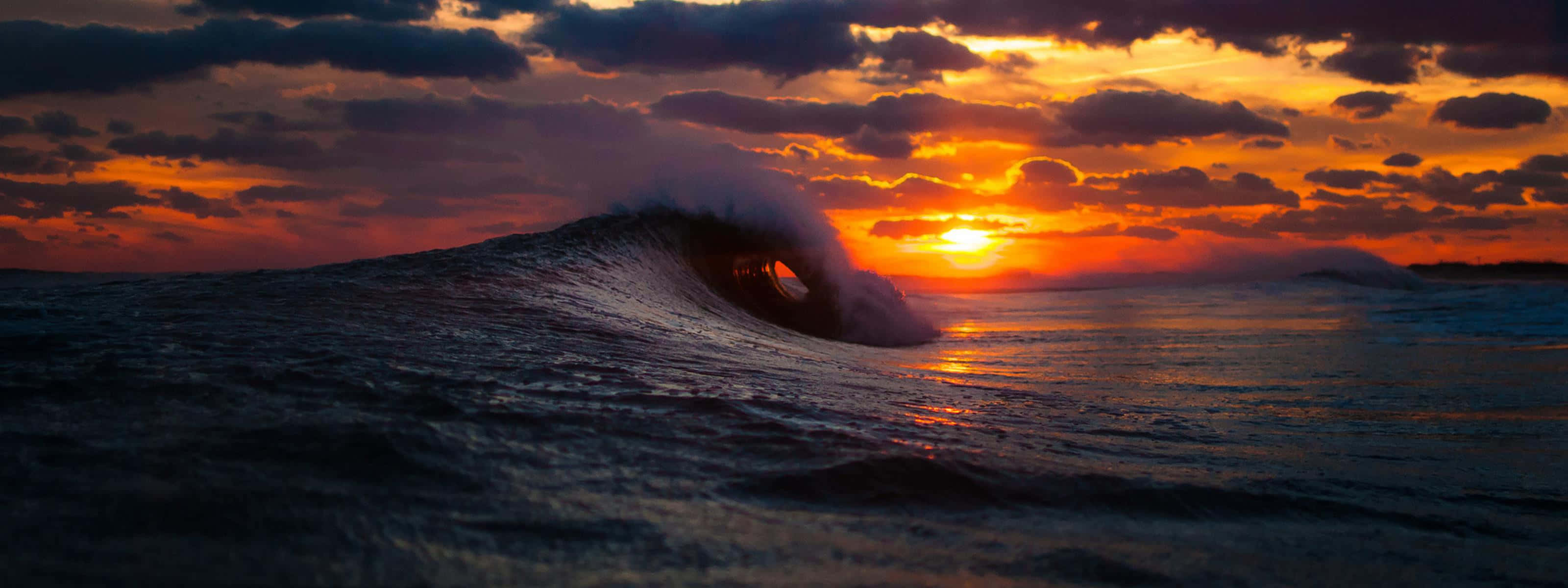 Sunset Wave 桌布 壁紙 — 巨大的海浪捲成完美的管狀，夕陽在其後落下，將火熱的橙色和紫色色調投射到天空和水面上。