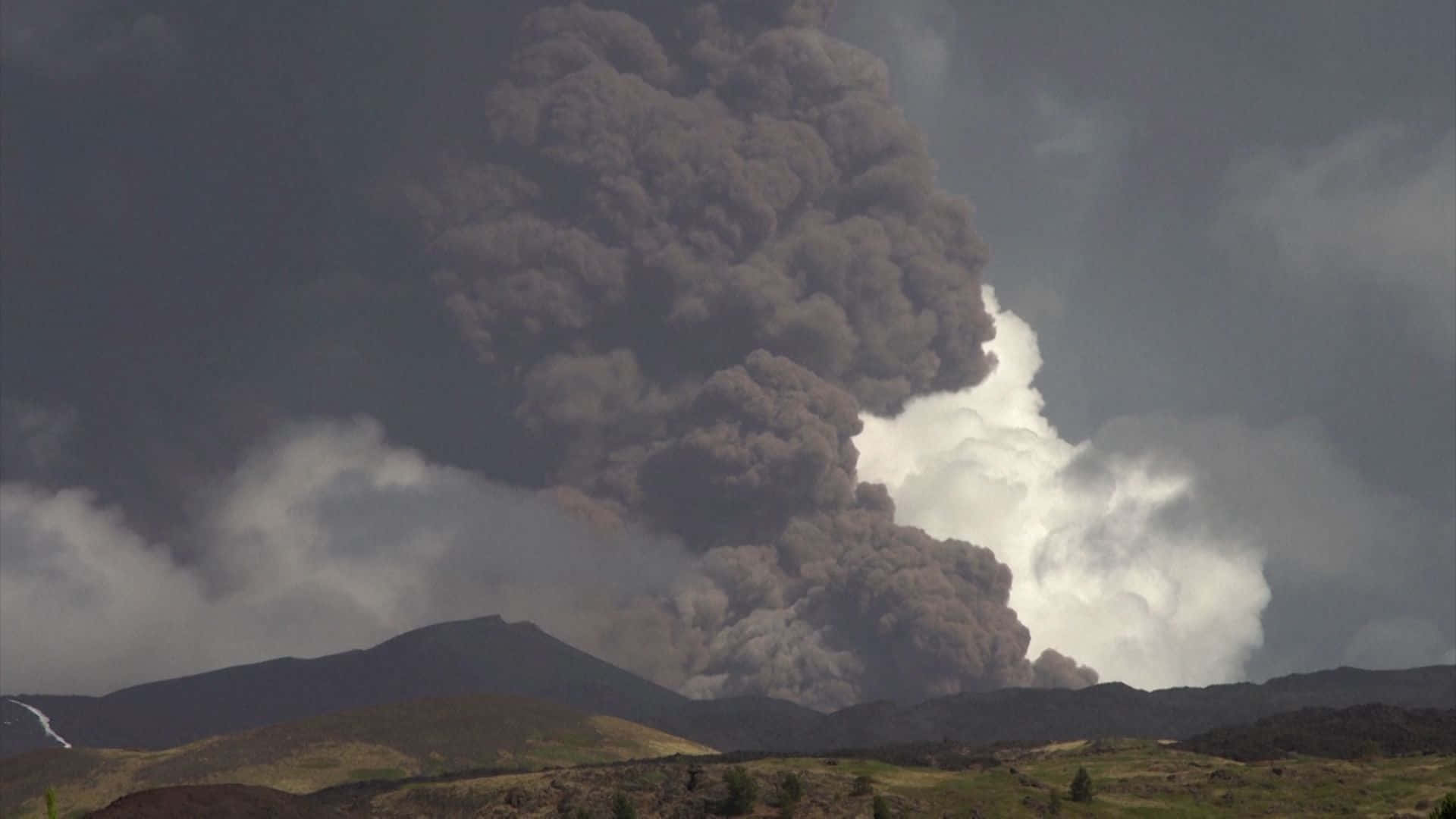 Volcano 桌布 壁紙 — 巨大的深色火山灰柱從火山噴發而出，背景是風暴天空和翻滾的雲層。
