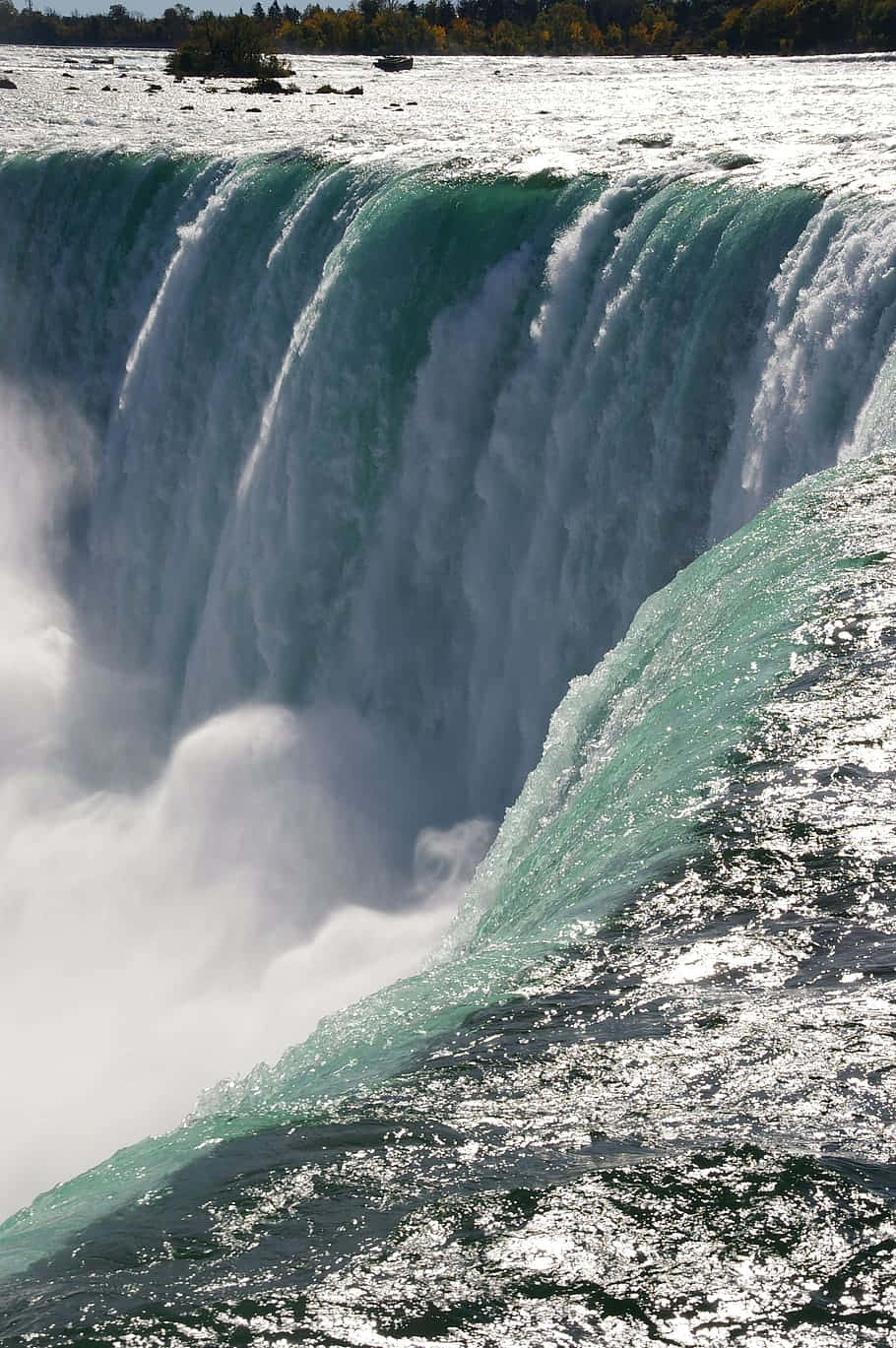 720p Niagara Falls 桌布 壁紙 — 巨大的瀑布傾瀉而下，碧綠色的水流激起霧氣，背景是秋日的樹木。
