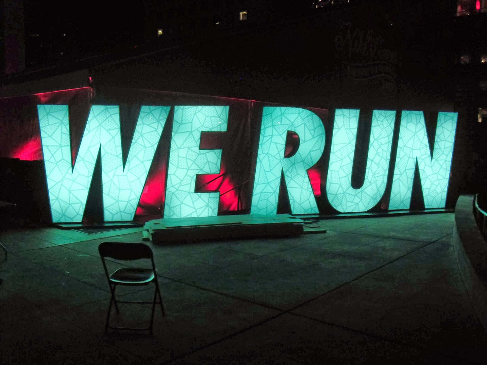 Run 桌布 壁紙 — 巨大的發光青綠色字母拼出WE RUN，在夜間以紅色背光照明，前景有一張孤獨的椅子。