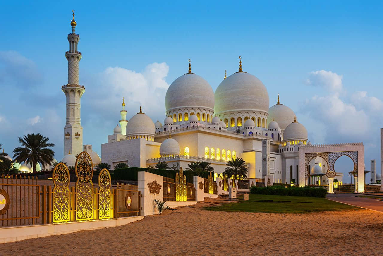 Uae 桌布 壁紙 — 巨大的白色清真寺，發光的圓頂和宣禮塔在黃昏時分照亮，背景是藍天和散落的雲朵。