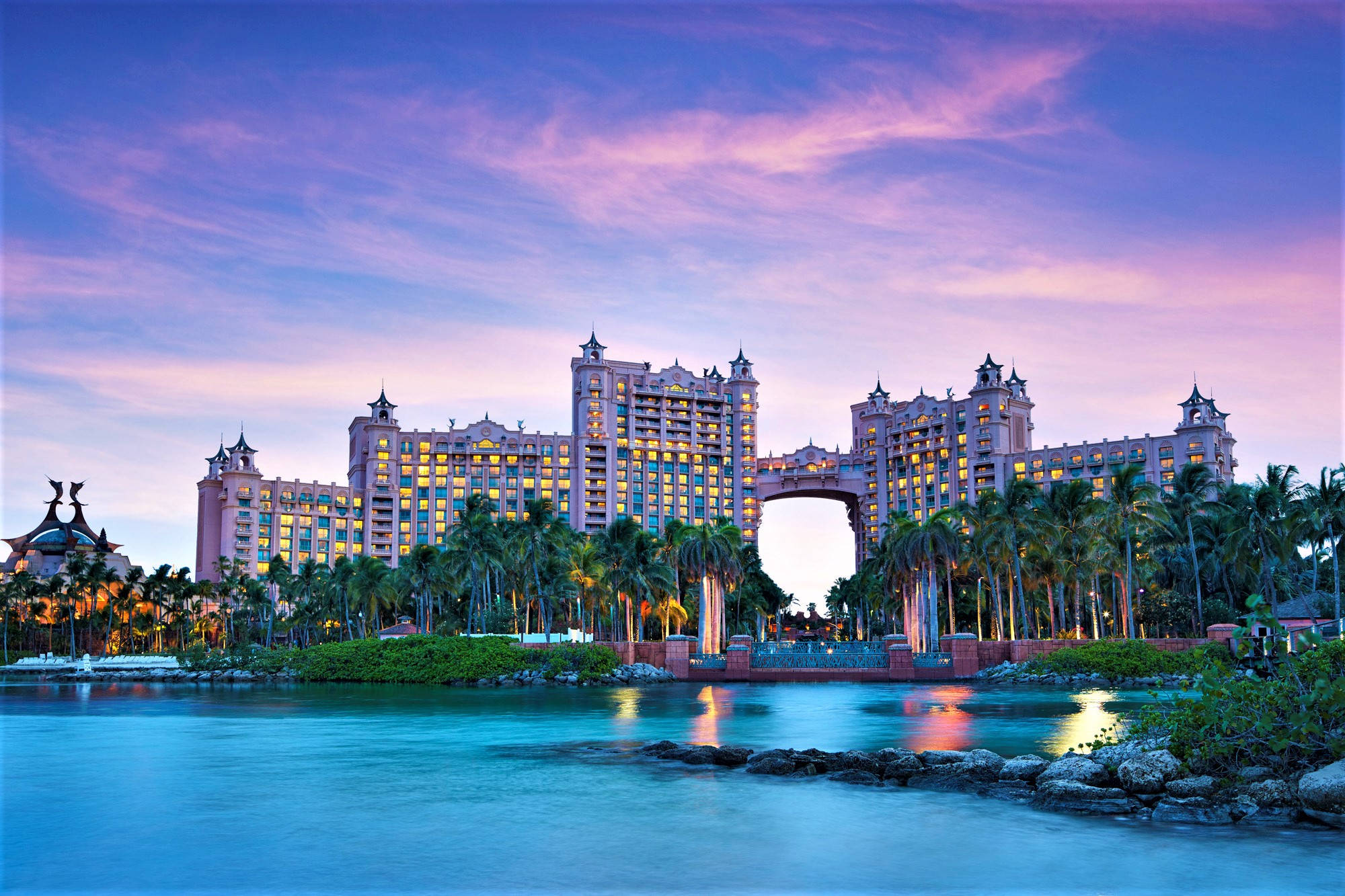 Nassau Bahamas 桌布 壁紙 — 巨大的粉紅色度假酒店，窗戶閃閃發光，在暮色中巍然聳立，棕櫚樹環繞，倒映在平靜的碧綠海水中。